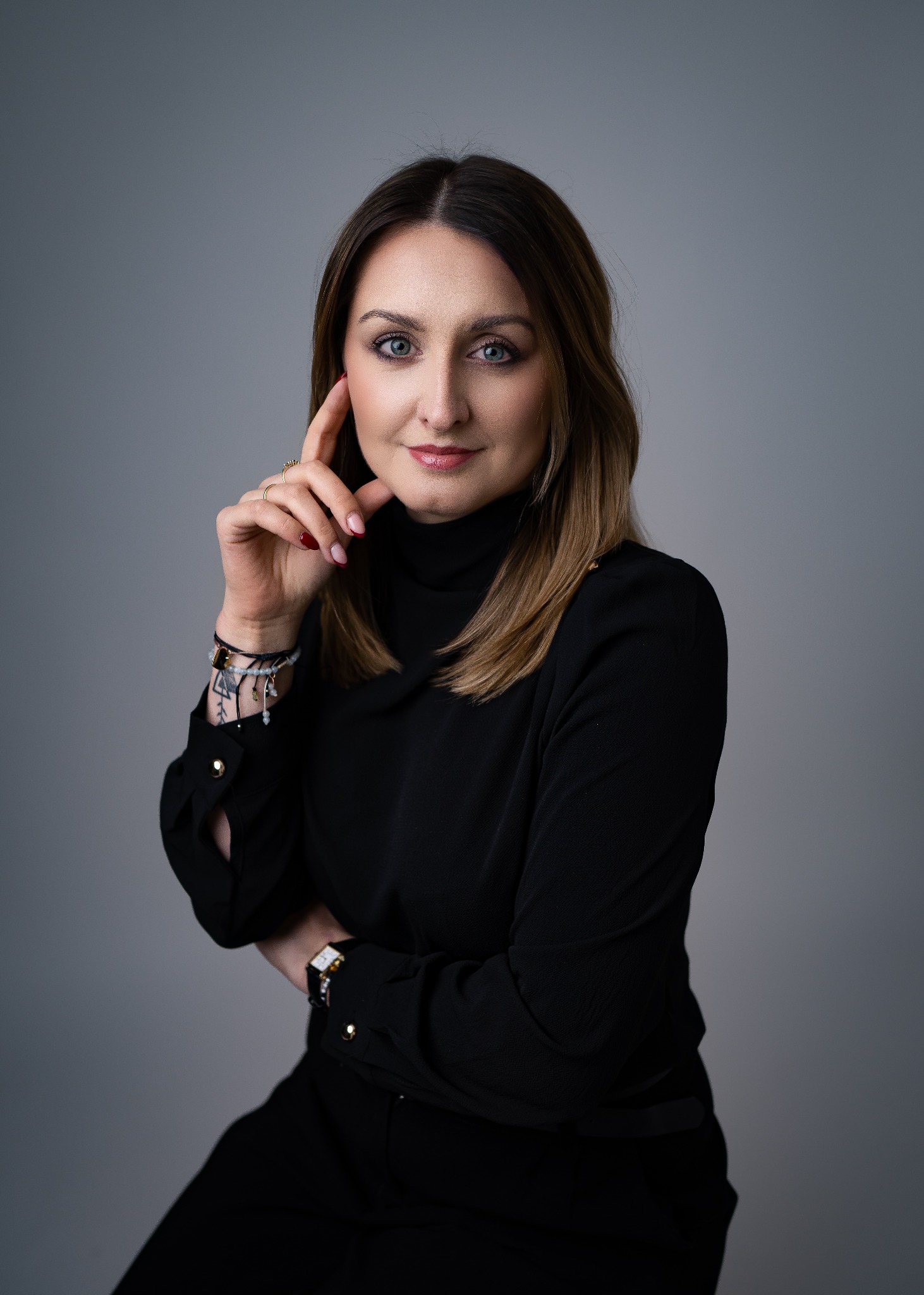 Portret kobiety w czarnej bluzce z golfem, z ręką przy twarzy, na szarym tle. Widoczny zegarek na nadgarstku i delikatna biżuteria.