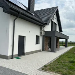 Nowoczesny dom jednorodzinny z białą elewacją, szarą stolarką okienną i dachem, panelami słonecznymi i dwoma klimatyzatorami na ścianie, widok z zewnątrz na kostkę brukową i trawnik.