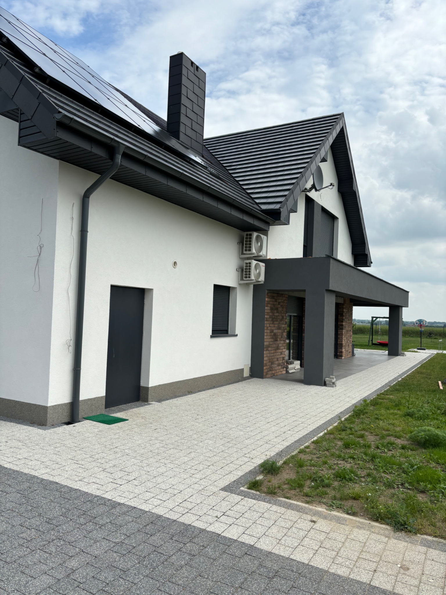 Nowoczesny dom jednorodzinny z białą elewacją, szarą stolarką okienną i dachem, panelami słonecznymi i dwoma klimatyzatorami na ścianie, widok z zewnątrz na kostkę brukową i trawnik.