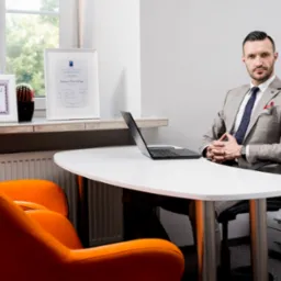 Mężczyzna w garniturze siedzi za biurkiem w biurze, obok okna z roślinami i certyfikatami, na biurku otwarty laptop.