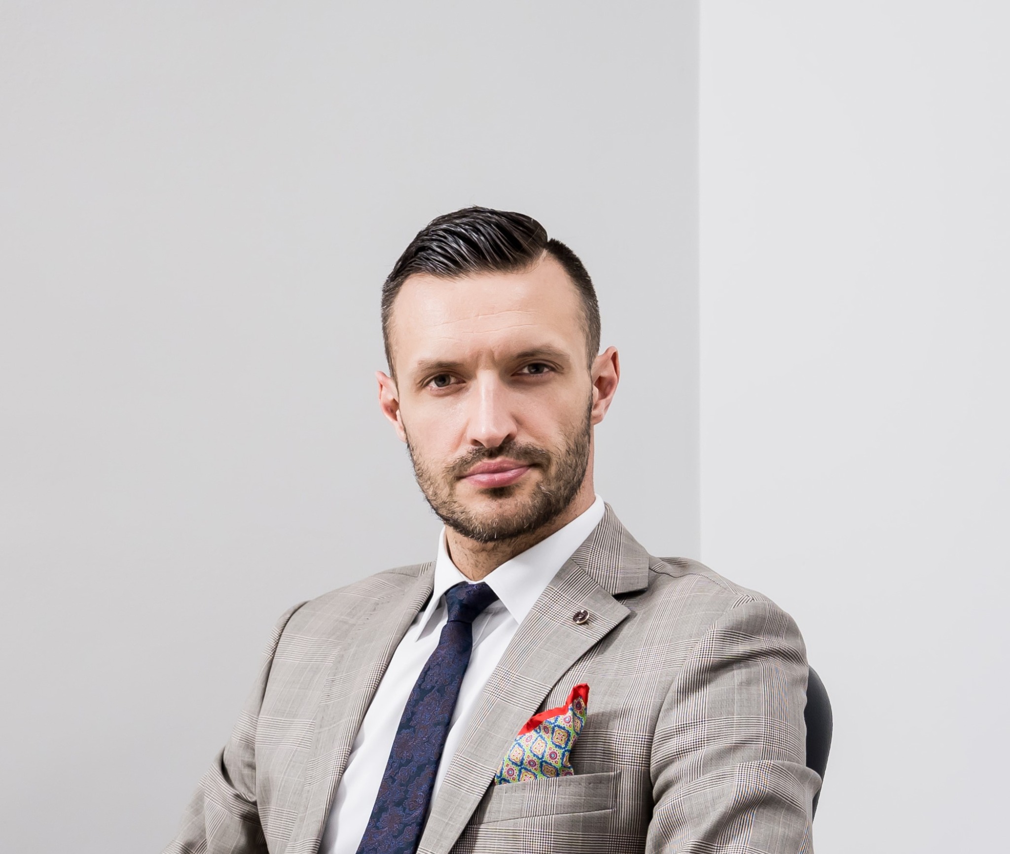 Mężczyzna w kraciastej marynarce, ciemnym krawacie i z wzorzystą poszetką w kieszonce, siedzący na krześle na tle jasnej ściany, patrzy w obiektyw.