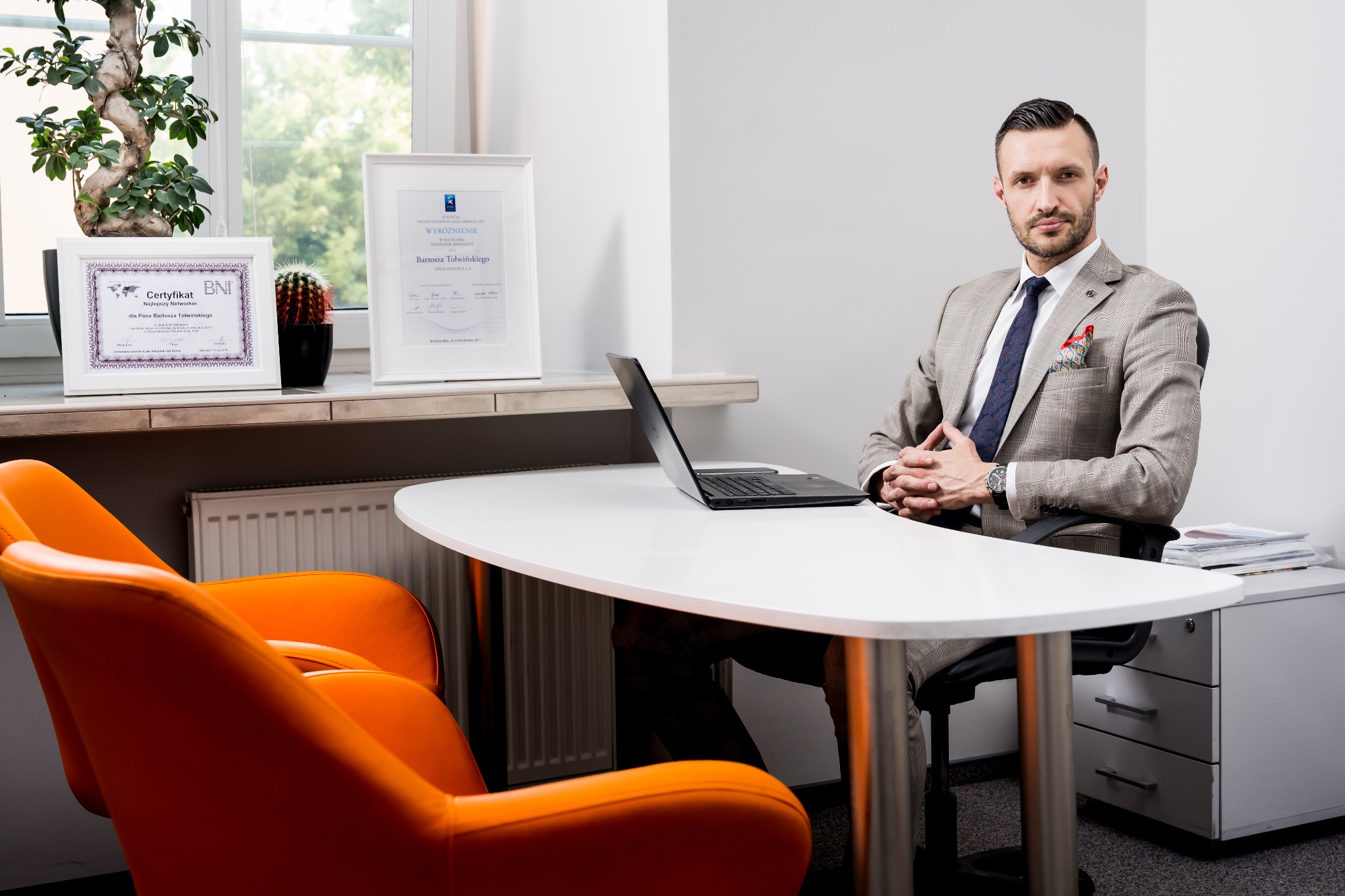 Mężczyzna w garniturze siedzi za biurkiem w biurze, obok okna z roślinami i certyfikatami, na biurku otwarty laptop.