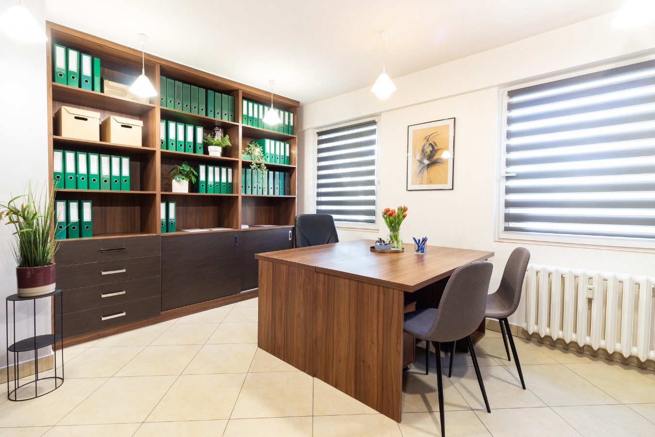 Jasne biuro z drewnianym regałem wypełnionym zielonymi segregatorami, biurkiem z tulipanami w wazonie i szarymi krzesłami, oświetlone lampami sufitowymi i naturalnym światłem z okien z żaluzjami...