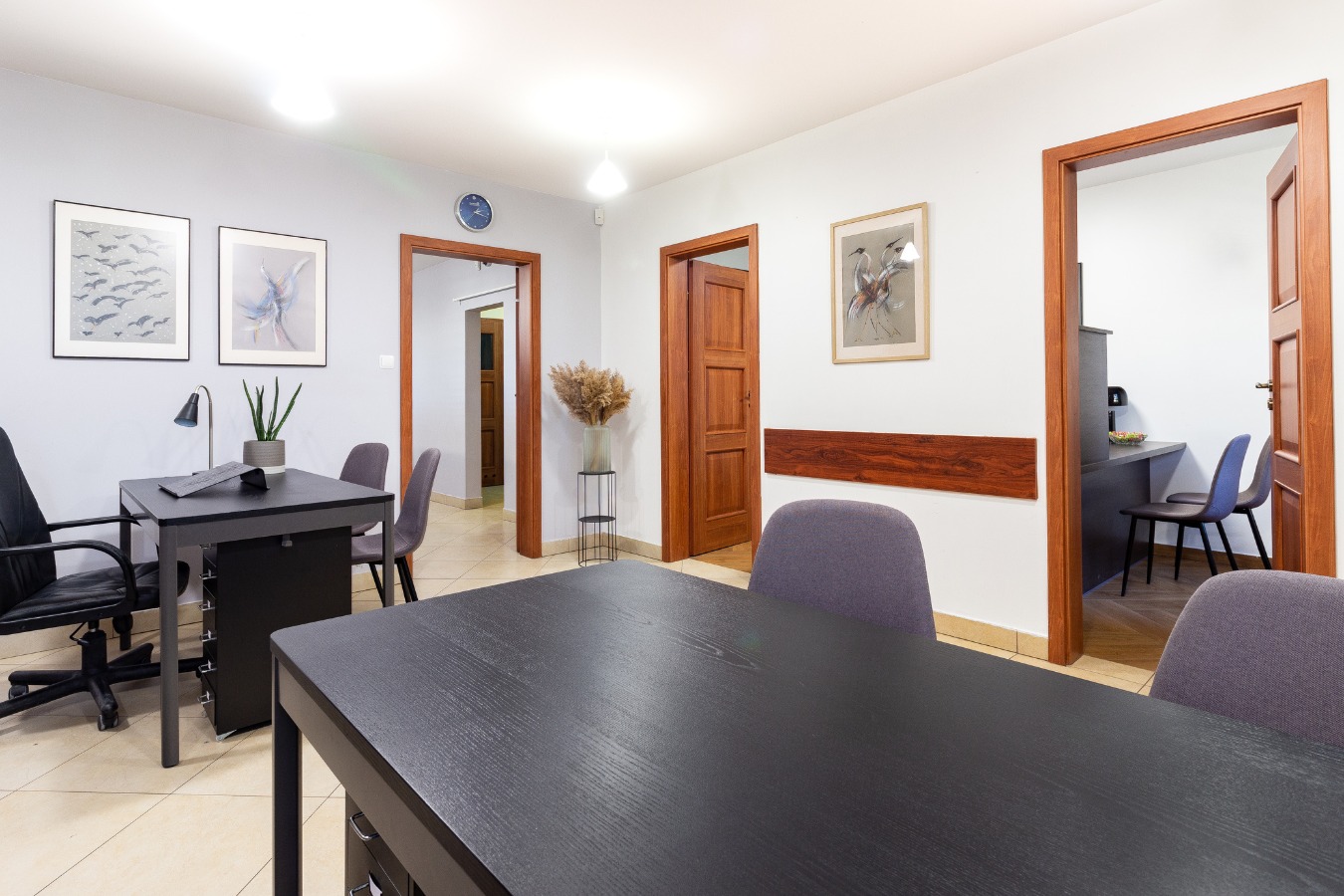 Jasne biuro w Białymstoku z dwoma biurkami, szarymi krzesłami, obrazami na ścianach i drewnianymi drzwiami, tworzące profesjonalne środowisko pracy.