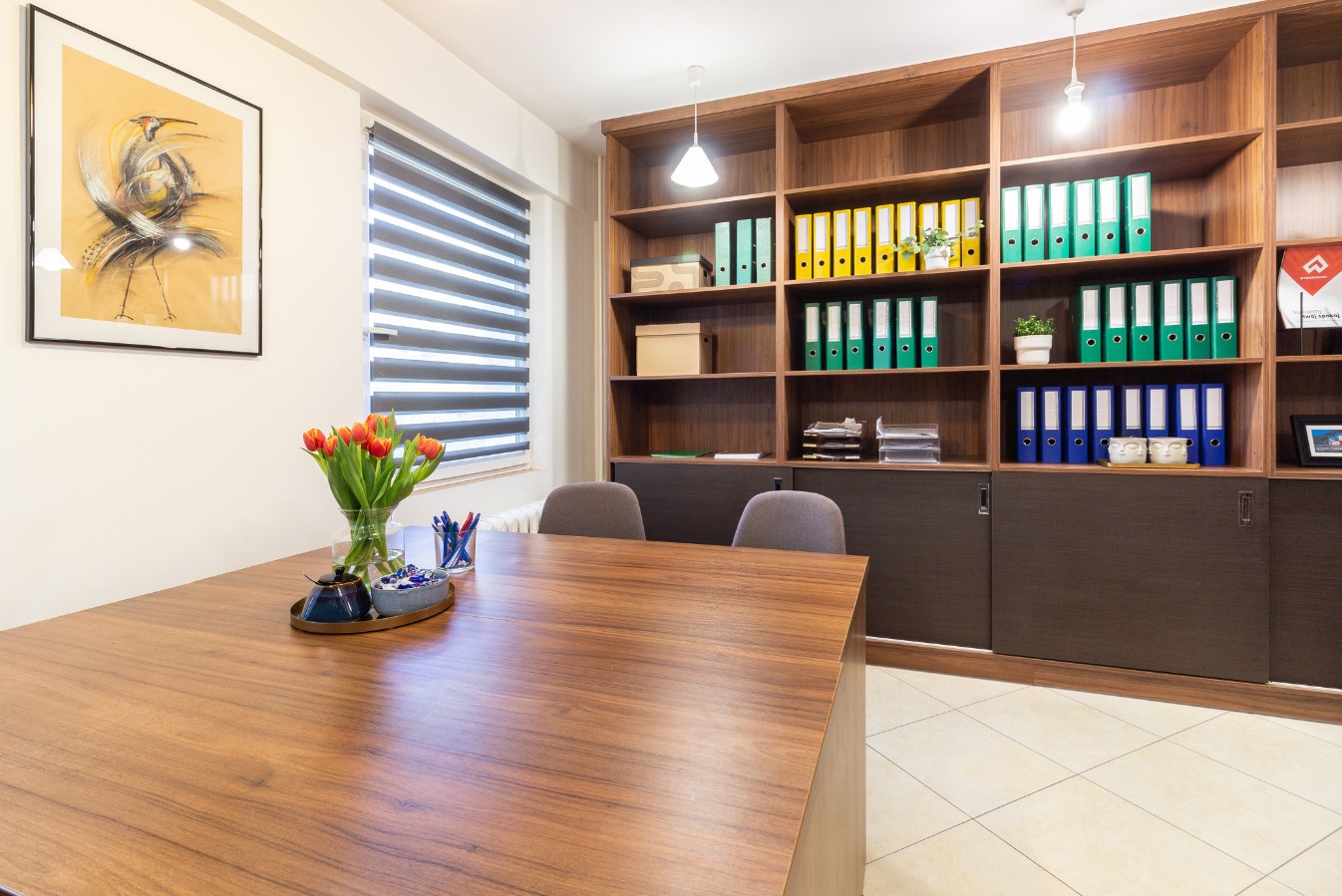 Biuro z drewnianym biurkiem, tulipanami w wazonie, oraz regałem wypełnionym segregatorami i dekoracjami, oświetlone lampą wiszącą.