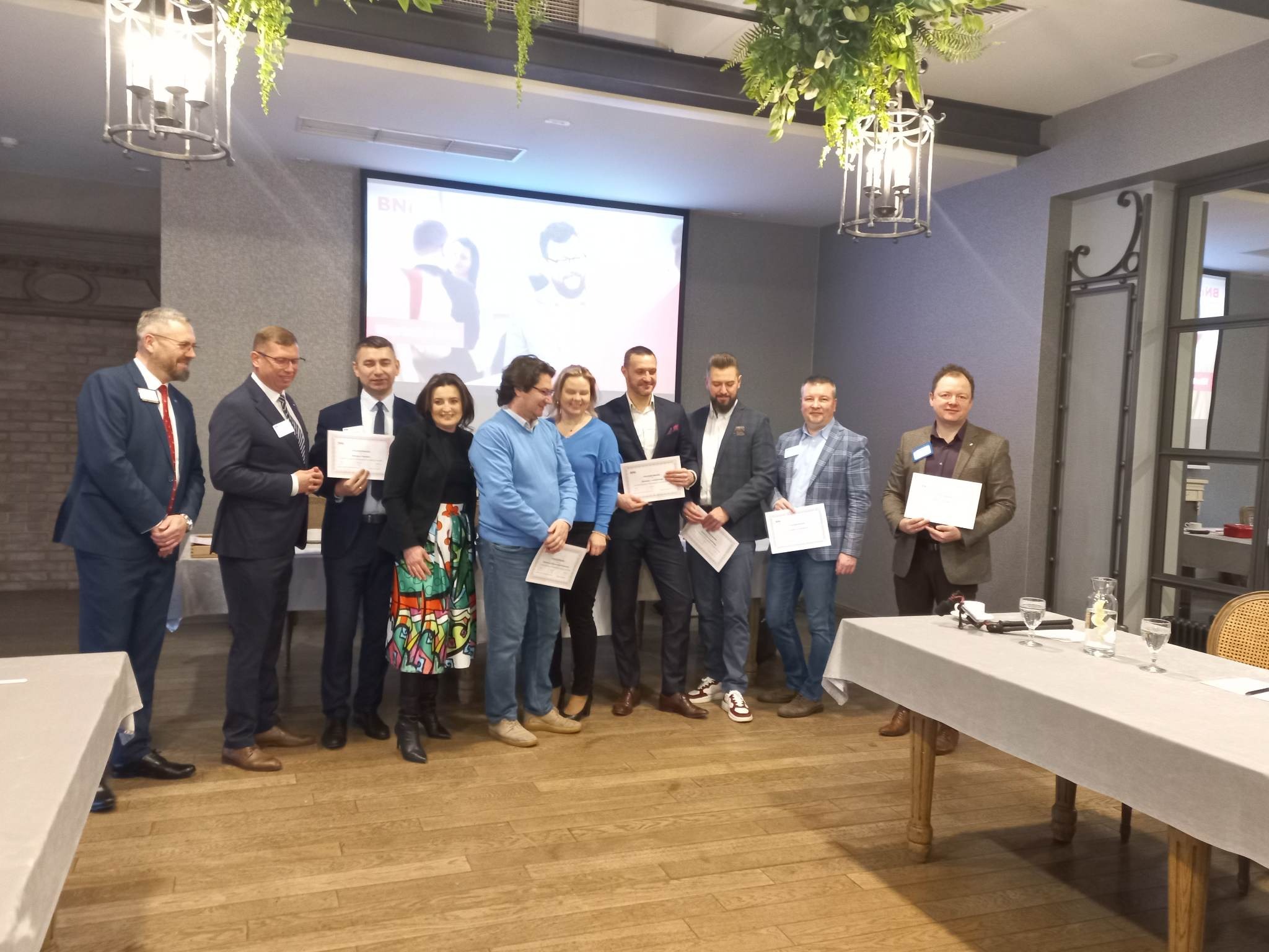 Grupa osób w formalnych strojach, stojąca na tle ekranu projekcyjnego i dekoracji roślinnych, trzymająca certyfikaty podczas wydarzenia networkingowego.