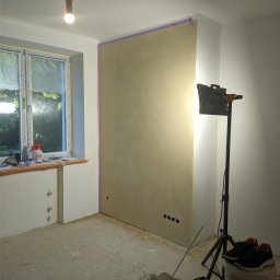 Luxs-Montaż-Bud - Pokój w trakcie prac wykończeniowych: ściana z nałożoną gładzią, zabezpieczona taśmą malarską, oświetlona lampą budowlaną na trójnogu, okno zakryte folią ochronną, widoczne narzędzia i puszki po...