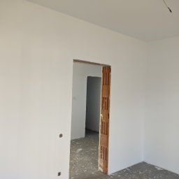 Luxs-Montaż-Bud - Wnętrze w trakcie prac wykończeniowych: świeżo pomalowane ściany na biało, surowa ościeżnica z cegły dziurawki, podłoga z resztkami zaprawy, widoczne gniazdka elektryczne.