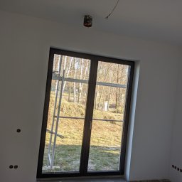 Luxs-Montaż-Bud - Wnętrze w trakcie remontu: białe ściany po szpachlowaniu i malowaniu, widoczne gniazdka elektryczne do wykończenia, okno z widokiem na zewnątrz, rusztowanie w tle, sufit z wystającym kablem.