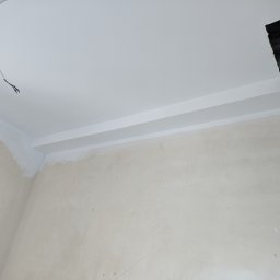 Luxs-Montaż-Bud - Pokój w trakcie prac wykończeniowych: surowa ściana, pomalowany na biało sufit z listwą dekoracyjną, wystający przewód elektryczny i otwór w suficie z widocznymi rurami.