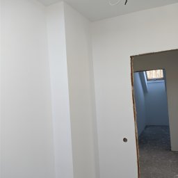 Luxs-Montaż-Bud - Świeżo pomalowane na biało wnętrze z widocznym otworem drzwiowym i wystającymi przewodami z sufitu.