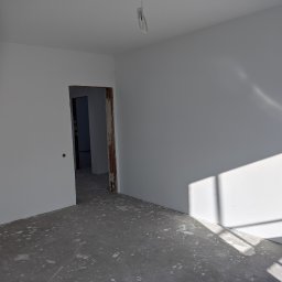 Luxs-Montaż-Bud - Świeżo pomalowane na biało wnętrze z widocznymi śladami farby na szarym, surowym betonie podłogi, z otwartym przejściem do kolejnego pomieszczenia i wiszącym przewodem oświetleniowym