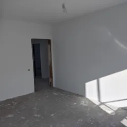 Świeżo pomalowane na biało wnętrze z widocznymi śladami farby na szarym, surowym betonie podłogi, z otwartym przejściem do kolejnego pomieszczenia i wiszącym przewodem oświetleniowym