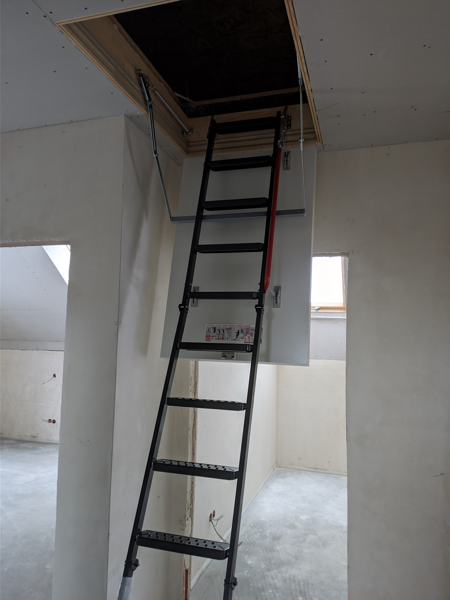 Rozkładana, czarna drabina strychowa z metalowymi stopniami, zamontowana w suficie z płyt kartonowo-gipsowych w trakcie prac wykończeniowych.