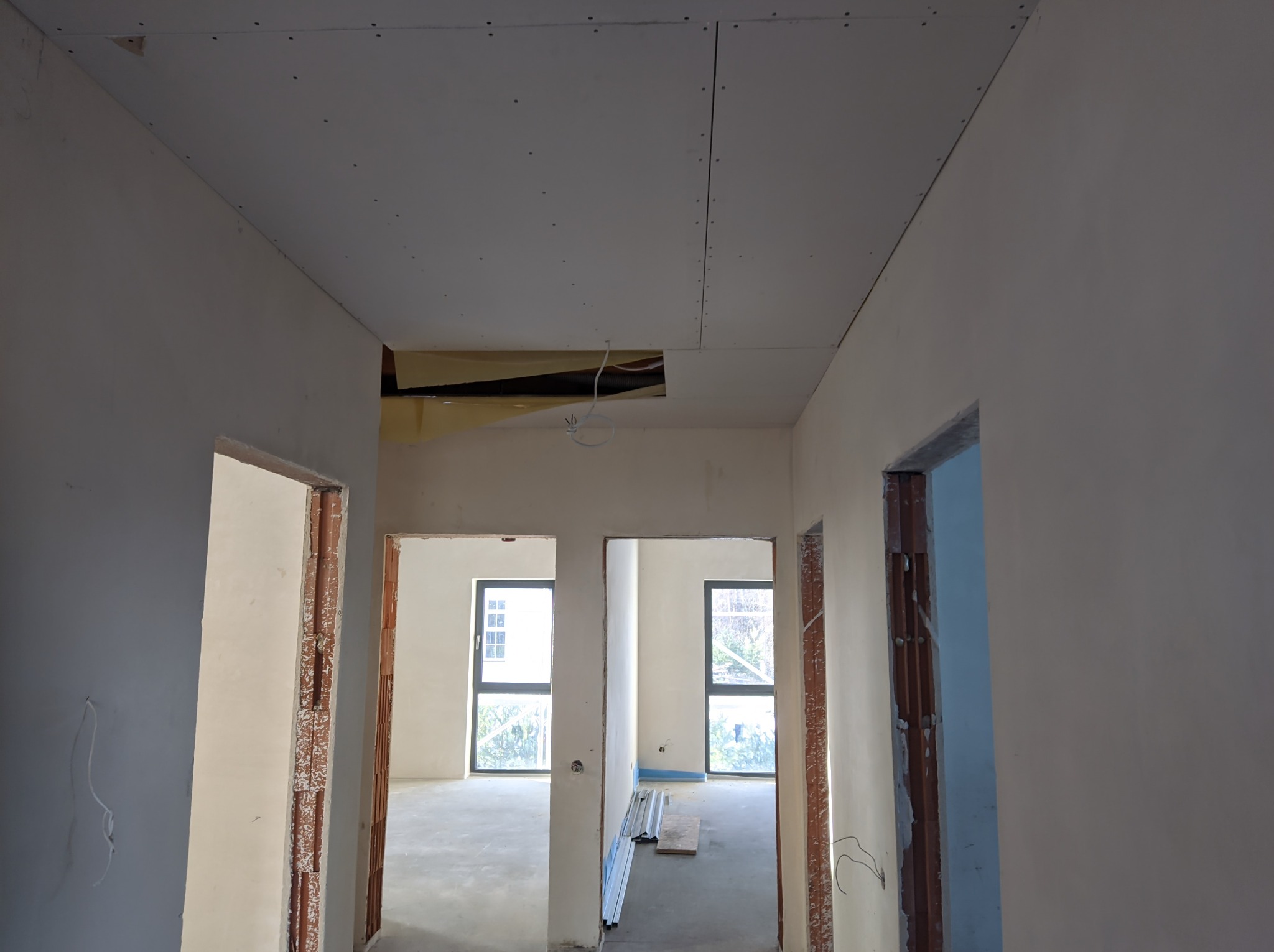 Wnętrze w trakcie prac wykończeniowych: widoczne ściany po szpachlowaniu, sufit z płyt kartonowo-gipsowych z widocznymi łączeniami i otworami na instalacje, oraz surowe ościeżnice drzwiowe.