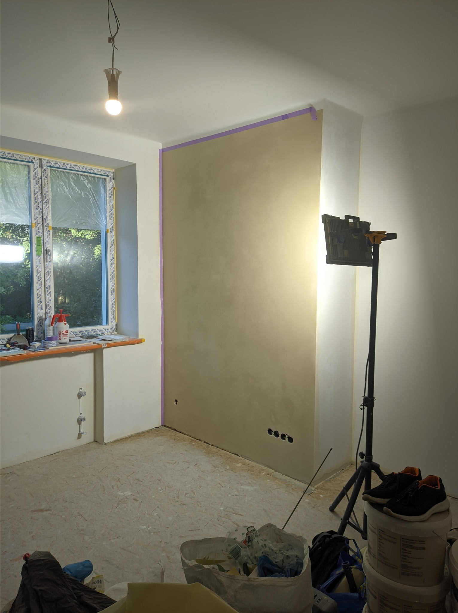 Pokój w trakcie prac wykończeniowych: ściana z nałożoną gładzią, zabezpieczona taśmą malarską, oświetlona lampą budowlaną na trójnogu, okno zakryte folią ochronną, widoczne narzędzia i puszki po...