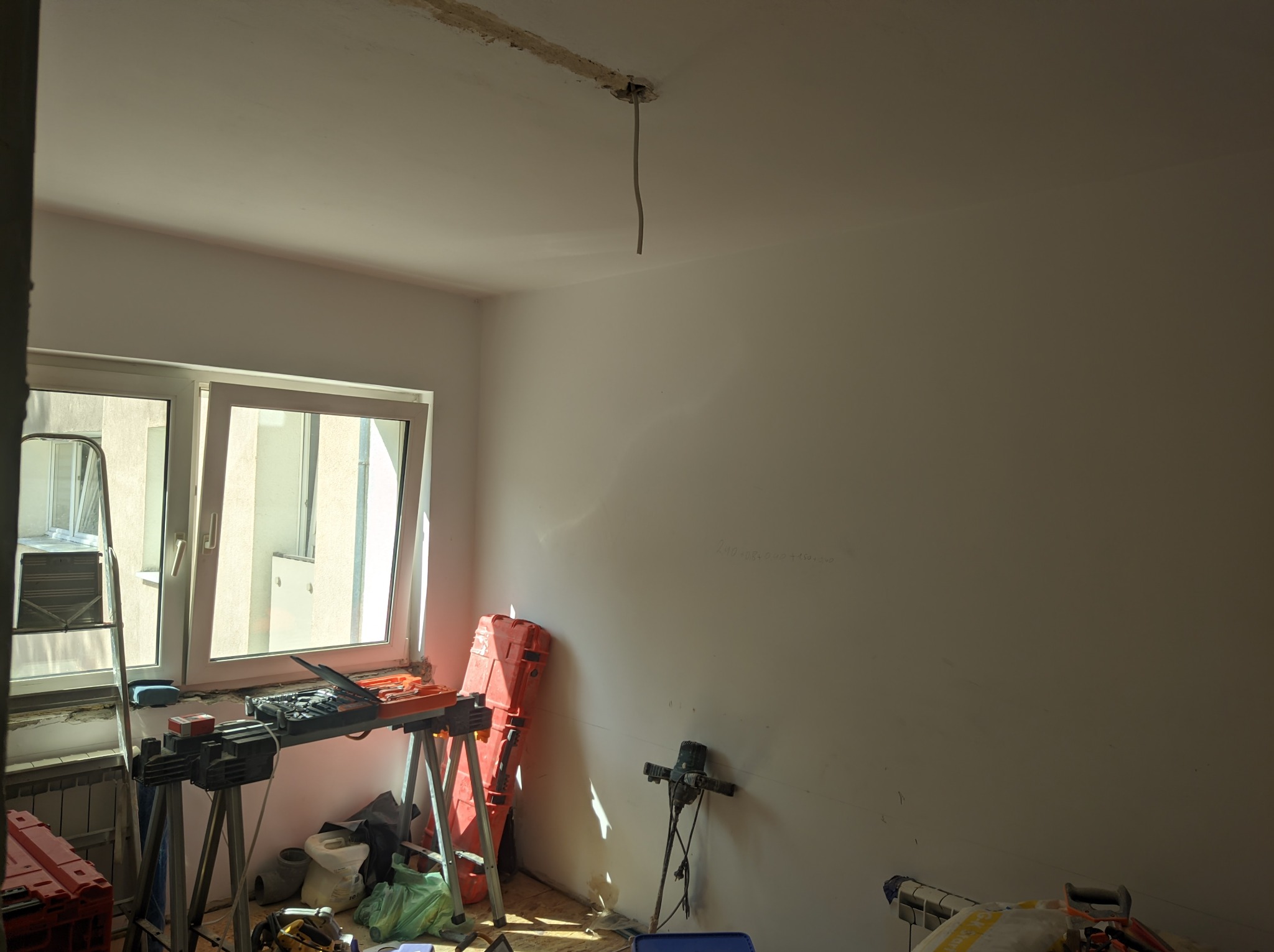 Pokój w trakcie przygotowań do prac wykończeniowych, z rozstawionymi narzędziami, drabina przy oknie, widoczne ślady po instalacji na suficie i notatki na ścianie.
