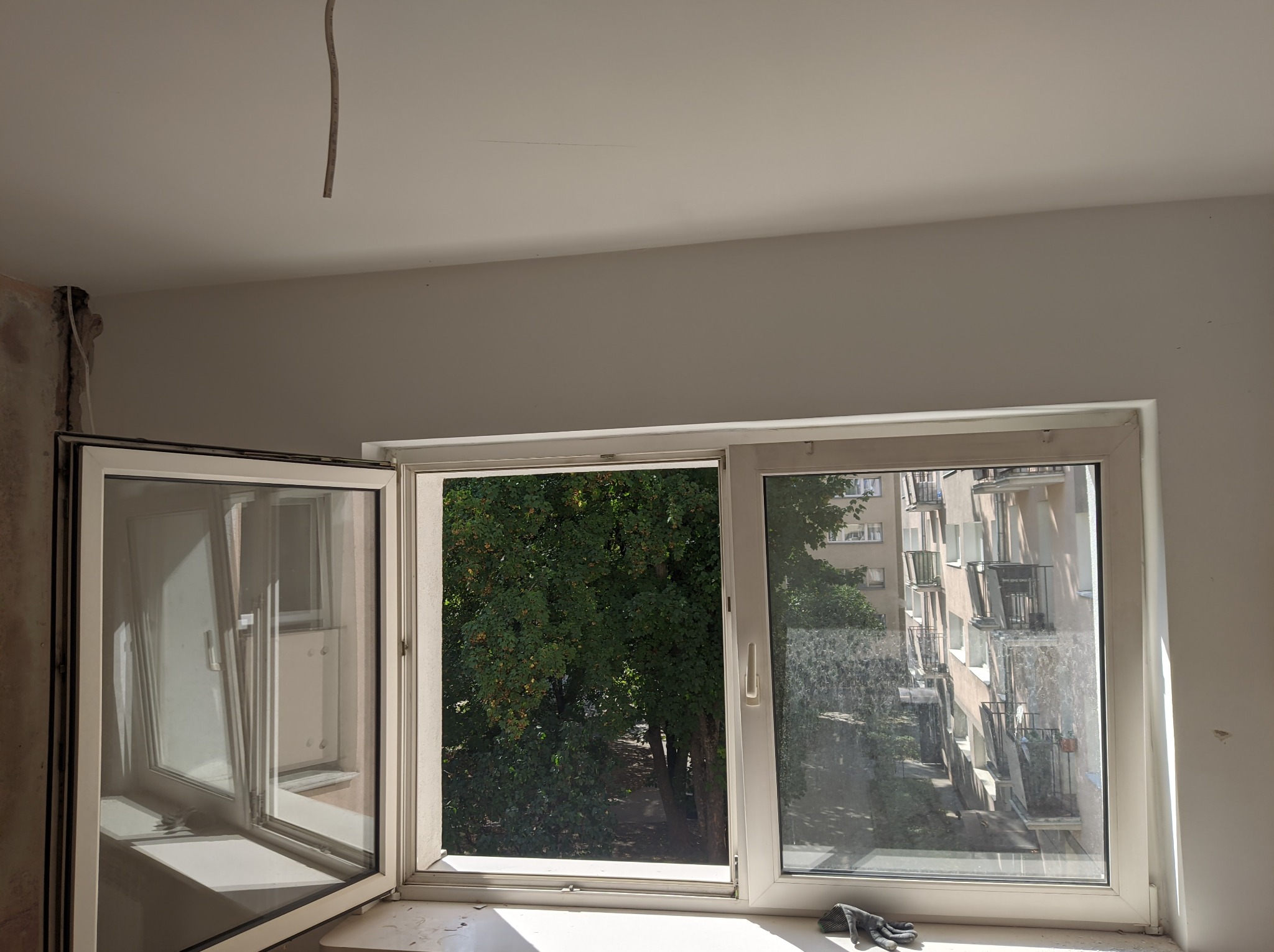 Wnętrze w trakcie remontu z otwartym oknem, widoczne ślady po instalacji elektrycznej na suficie i niedokończone wykończenie ściany obok okna.