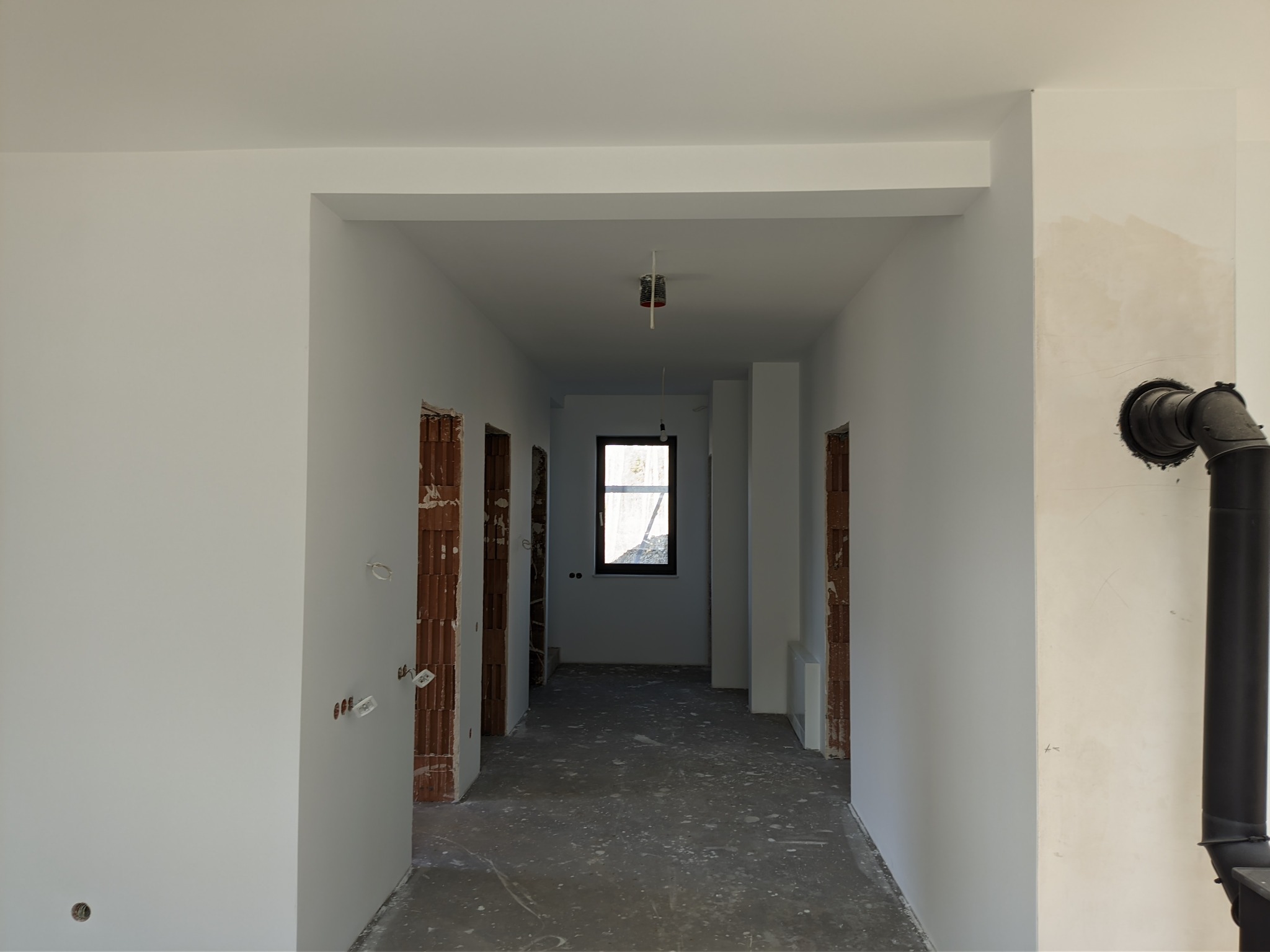Korytarz w trakcie remontu: ściany po szpachlowaniu i malowaniu na biało, widoczne instalacje elektryczne i rury wentylacyjne, surowy beton na podłodze, niezabezpieczone otwory drzwiowe.