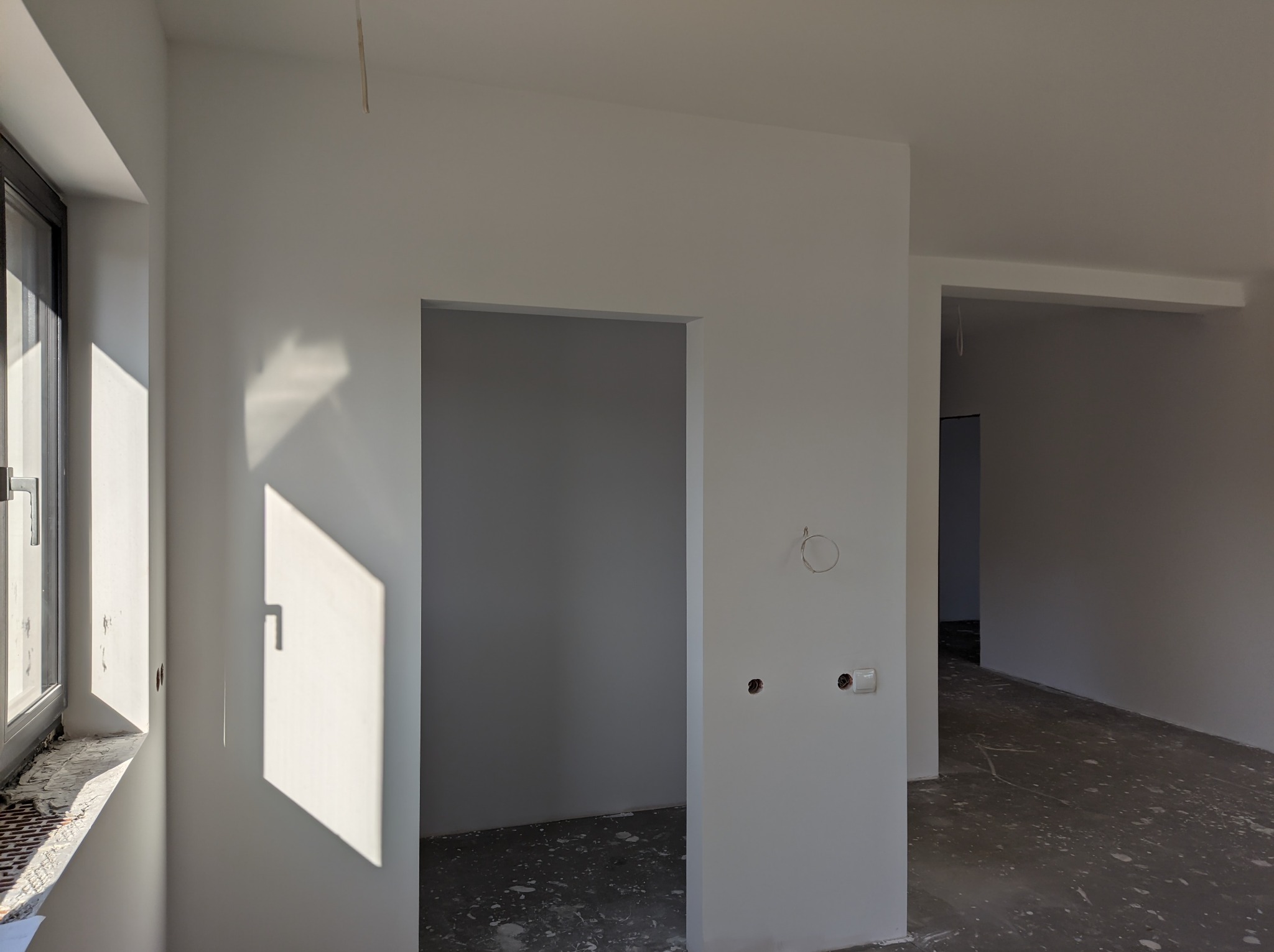 Wnętrze w trakcie wykańczania: białe ściany, wpadające przez okno promienie słoneczne, widoczne otwory na gniazdka elektryczne i niedokończona podłoga.