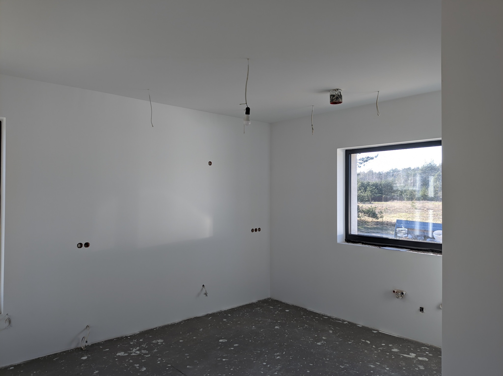 Pomalowane na biało wnętrze pokoju z widocznymi puszkami instalacyjnymi, zwisającymi kablami i surową podłogą, widok z okna na zewnątrz.