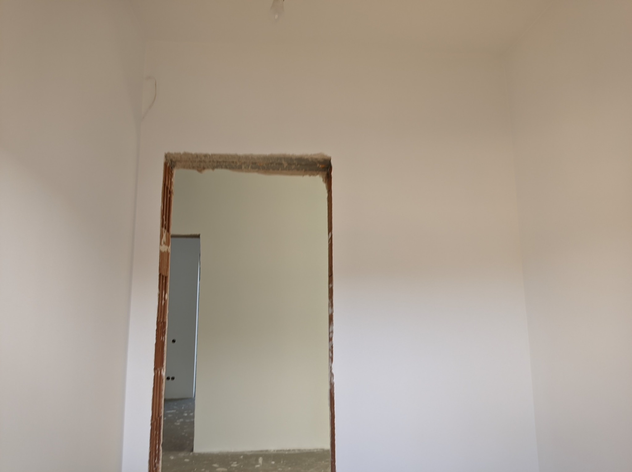 Wnętrze pokoju w trakcie remontu, widoczne świeżo pomalowane ściany na biało i surowy otwór drzwiowy z ceglaną ościeżnicą, w tle widoczne kolejne pomieszczenie.