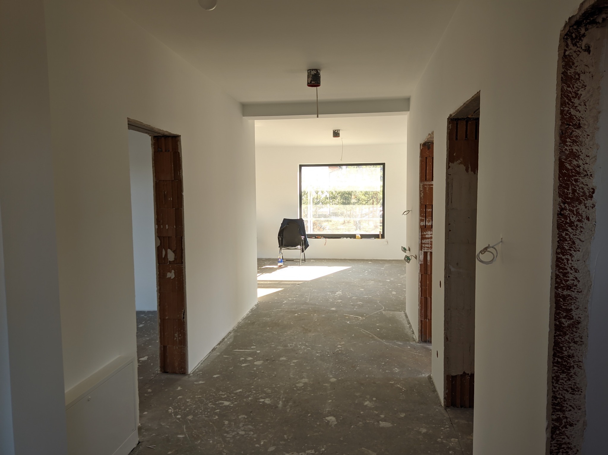 Korytarz w trakcie prac wykończeniowych: świeżo pomalowane, białe ściany, surowa podłoga, widoczne fragmenty cegieł wokół ościeżnic, w głębi pokoju krzesło pod oknem.
