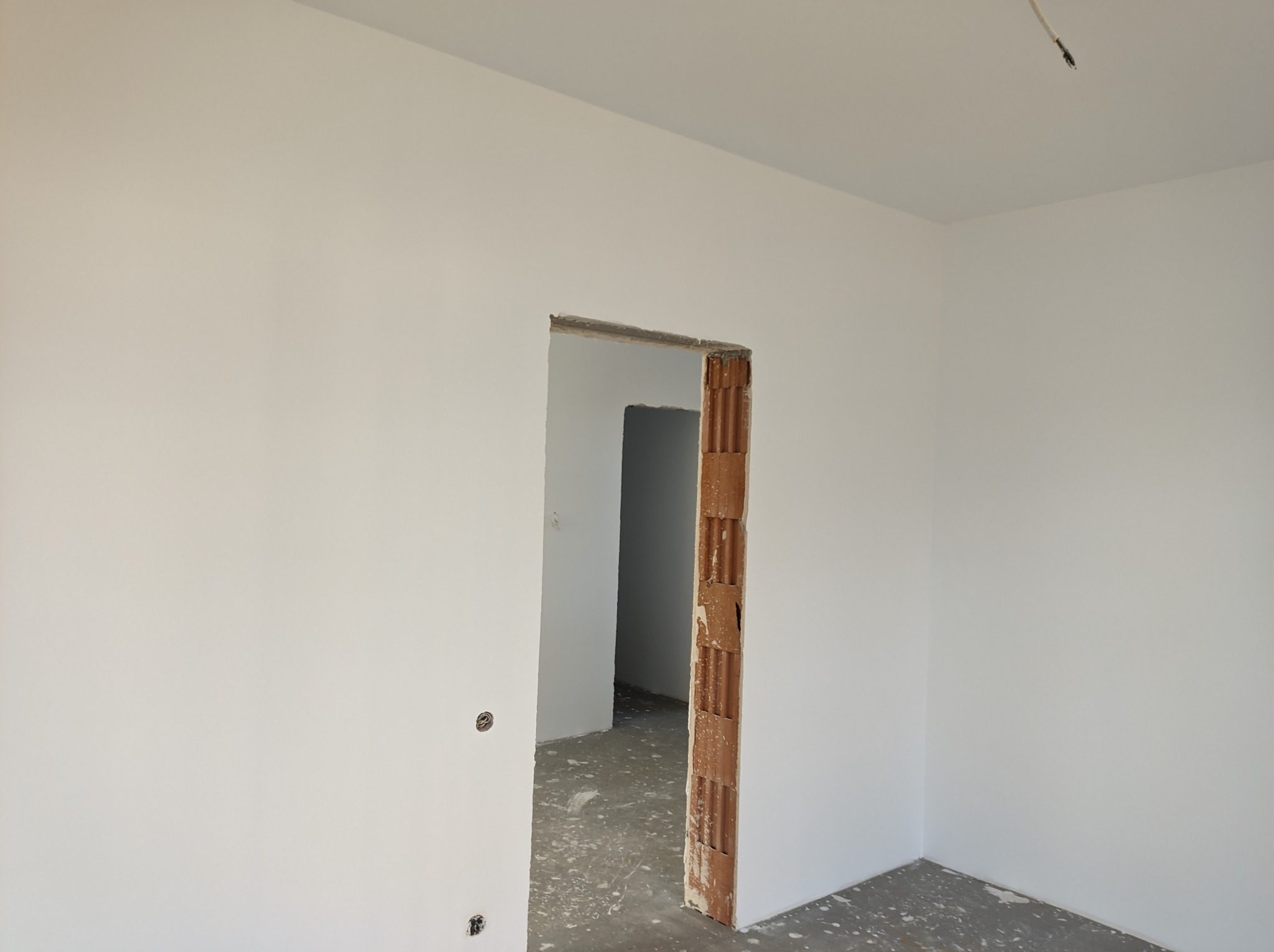 Wnętrze w trakcie prac wykończeniowych: świeżo pomalowane ściany na biało, surowa ościeżnica z cegły dziurawki, podłoga z resztkami zaprawy, widoczne gniazdka elektryczne.