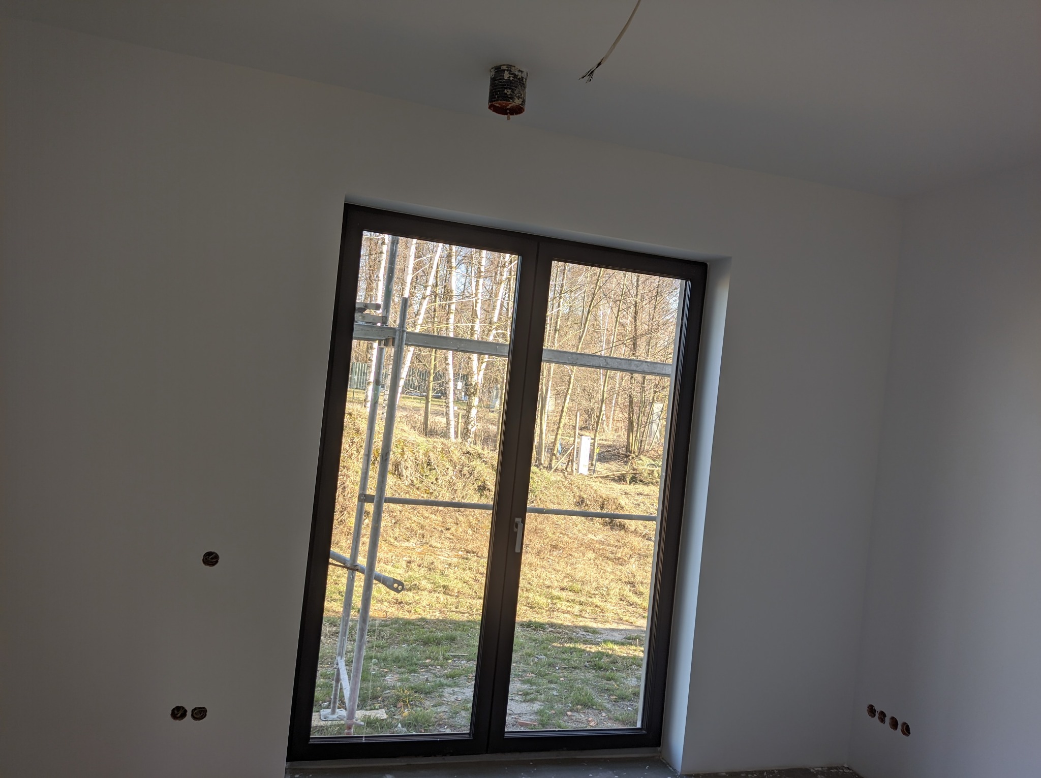 Wnętrze w trakcie remontu: białe ściany po szpachlowaniu i malowaniu, widoczne gniazdka elektryczne do wykończenia, okno z widokiem na zewnątrz, rusztowanie w tle, sufit z wystającym kablem.