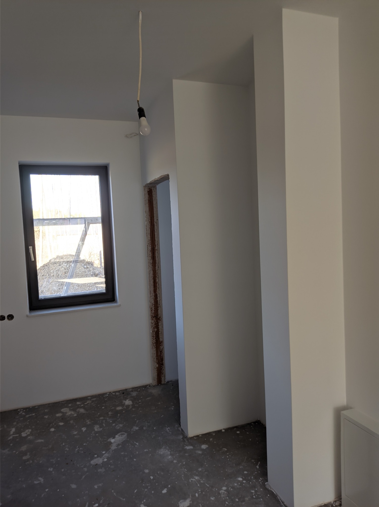 Wnętrze w trakcie prac, świeżo pomalowane ściany na biało, surowy beton na podłodze z widocznymi plamami farby, wisząca żarówka na kablu, surowa ościeżnica drzwiowa.