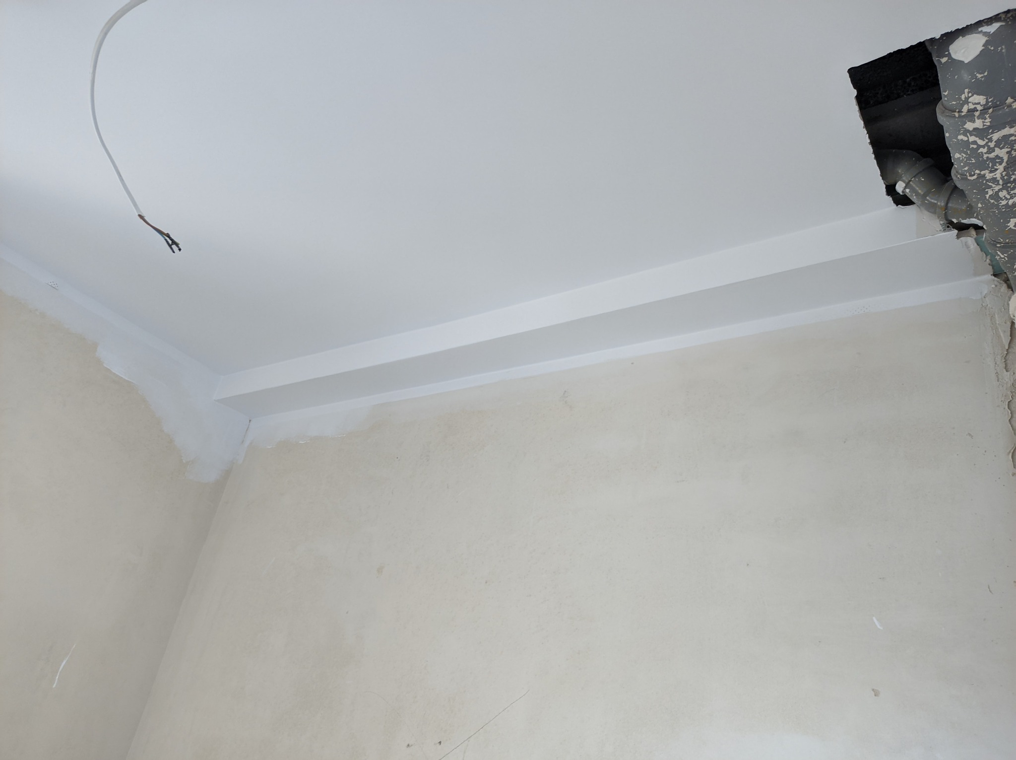 Pokój w trakcie prac wykończeniowych: surowa ściana, pomalowany na biało sufit z listwą dekoracyjną, wystający przewód elektryczny i otwór w suficie z widocznymi rurami.