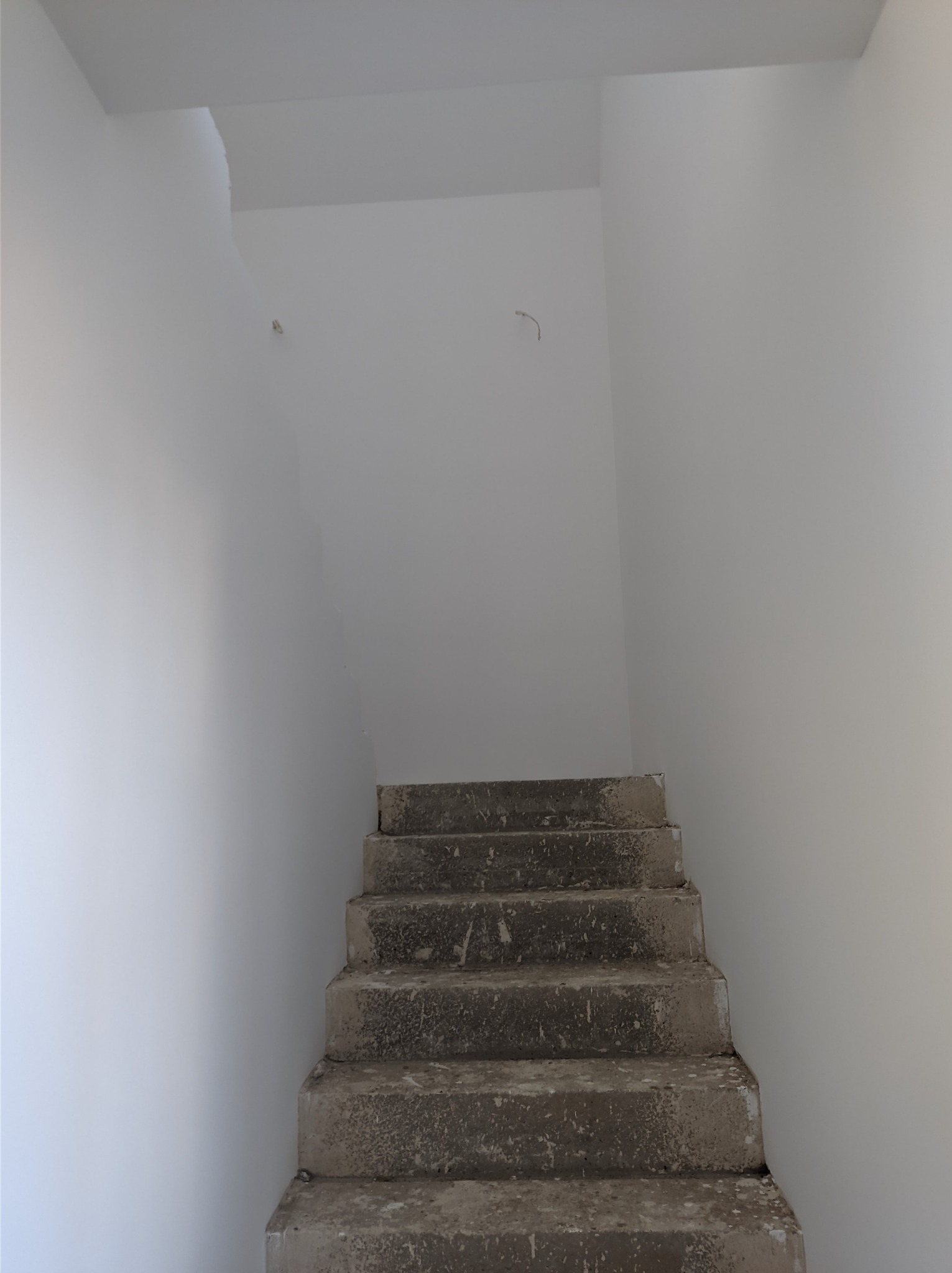 Surowe, betonowe schody wewnątrz budynku z pomalowanymi na biało ścianami, widoczne niedociągnięcia w górnej części ściany i wystający kabel.