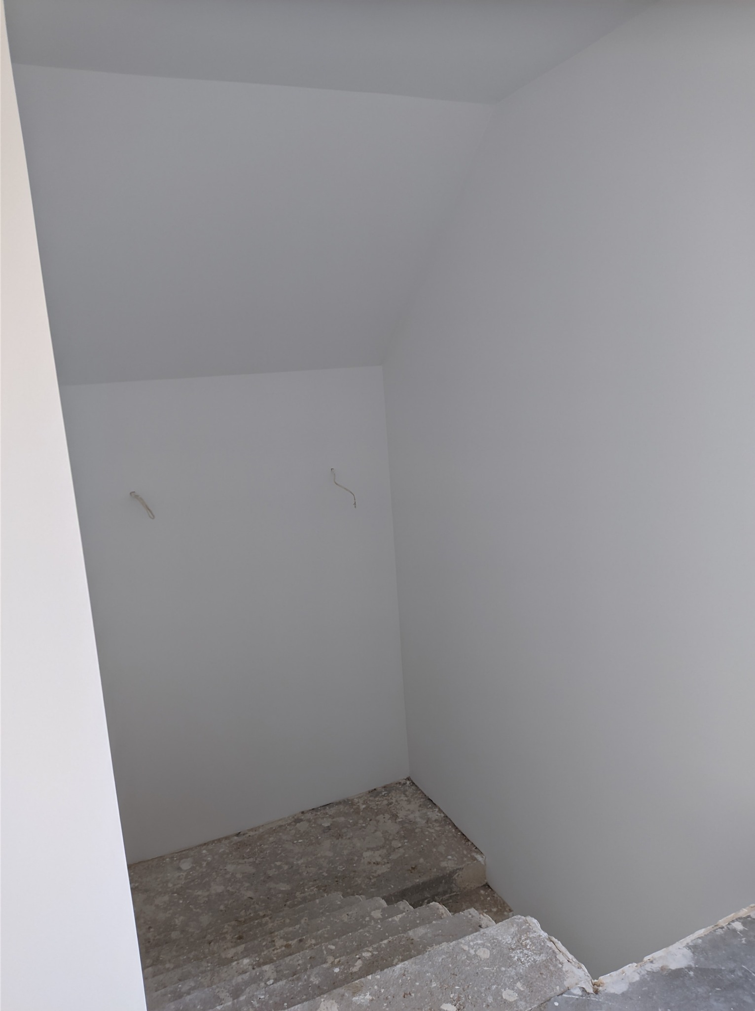 Świeżo pomalowane, jasnoszare ściany klatki schodowej w trakcie prac wykończeniowych, widoczne niedoskonałości na schodach i wystające kable ze ściany.