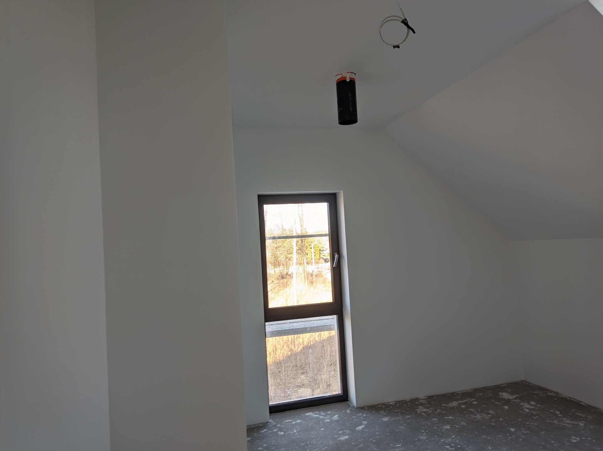 Świeżo wykończone poddasze z oknem, białe ściany i szary betonowy podkład podłogowy, widoczna instalacja elektryczna na suficie.