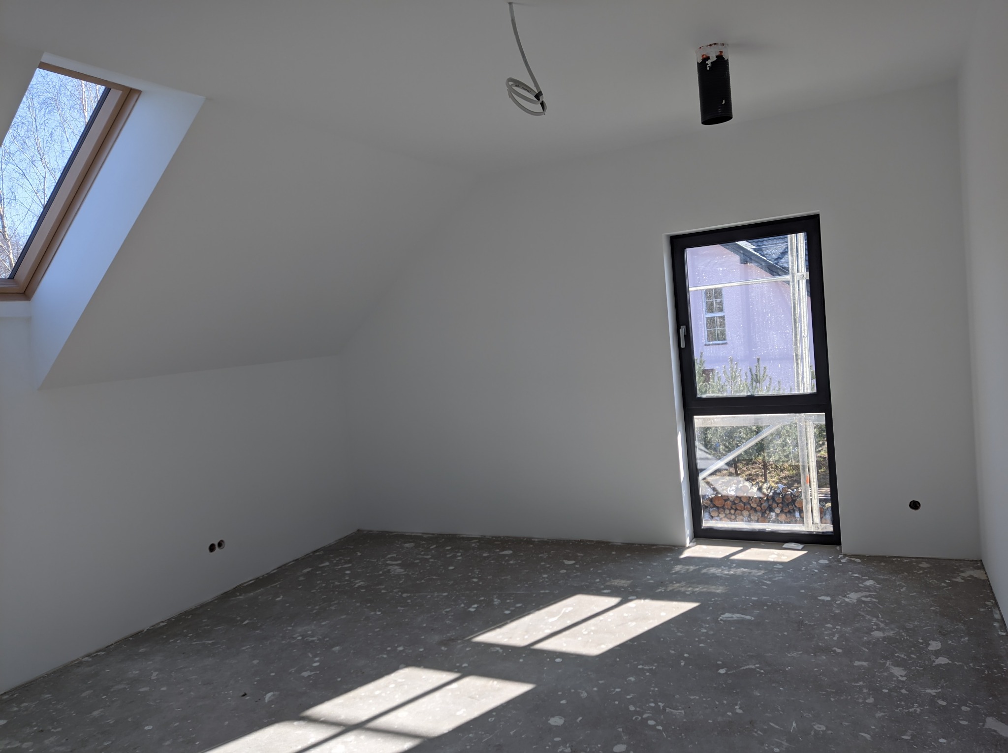 Pokój w stanie deweloperskim z oknem dachowym i pionowym, surowa podłoga z widocznymi śladami po pracach wykończeniowych, białe ściany z wystającymi przewodami elektrycznymi.
