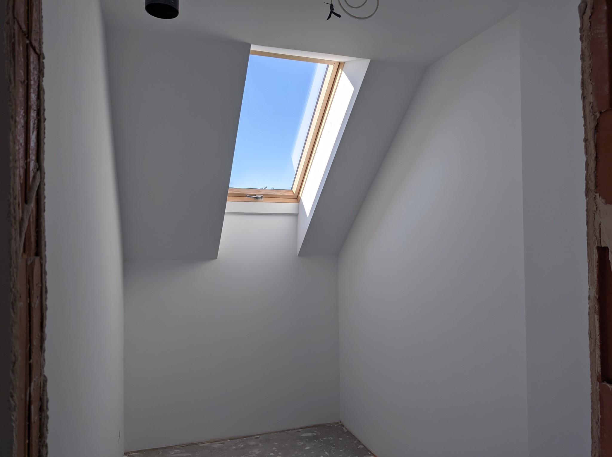 Poddasze w trakcie wykańczania, widoczne białe ściany, okno dachowe z widokiem na błękitne niebo, surowy fragment muru z cegły po lewej stronie i betonowa podłoga w szarym odcieniu.