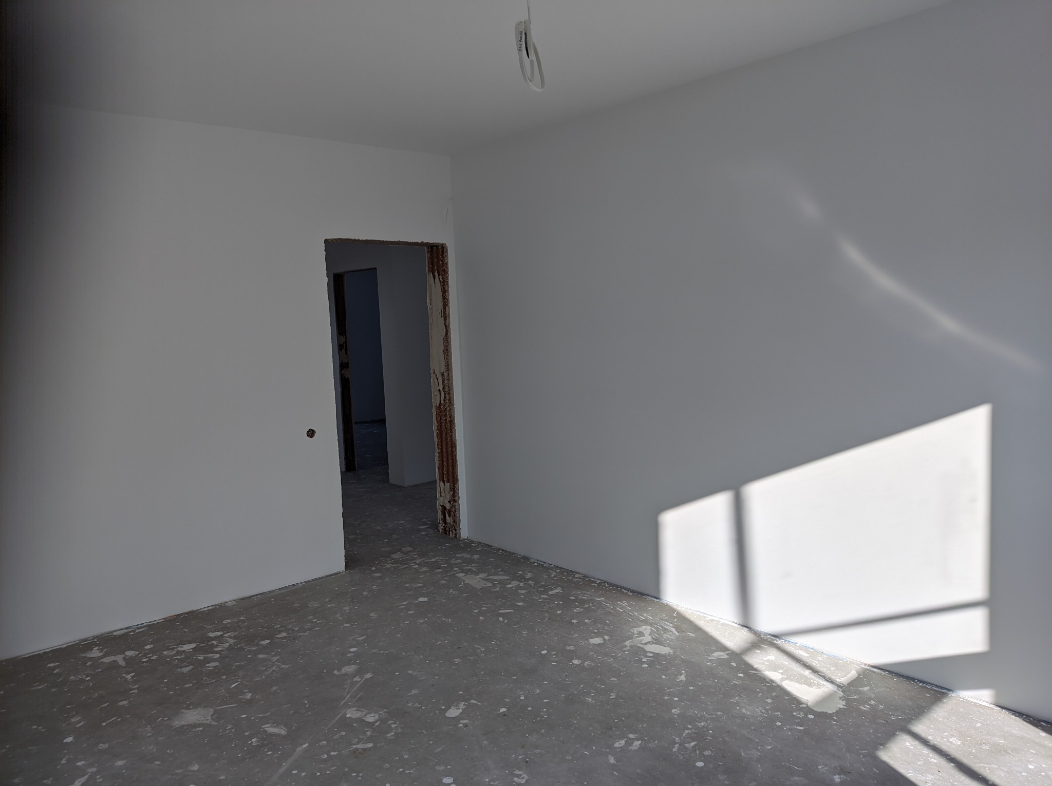 Świeżo pomalowane na biało wnętrze z widocznymi śladami farby na szarym, surowym betonie podłogi, z otwartym przejściem do kolejnego pomieszczenia i wiszącym przewodem oświetleniowym