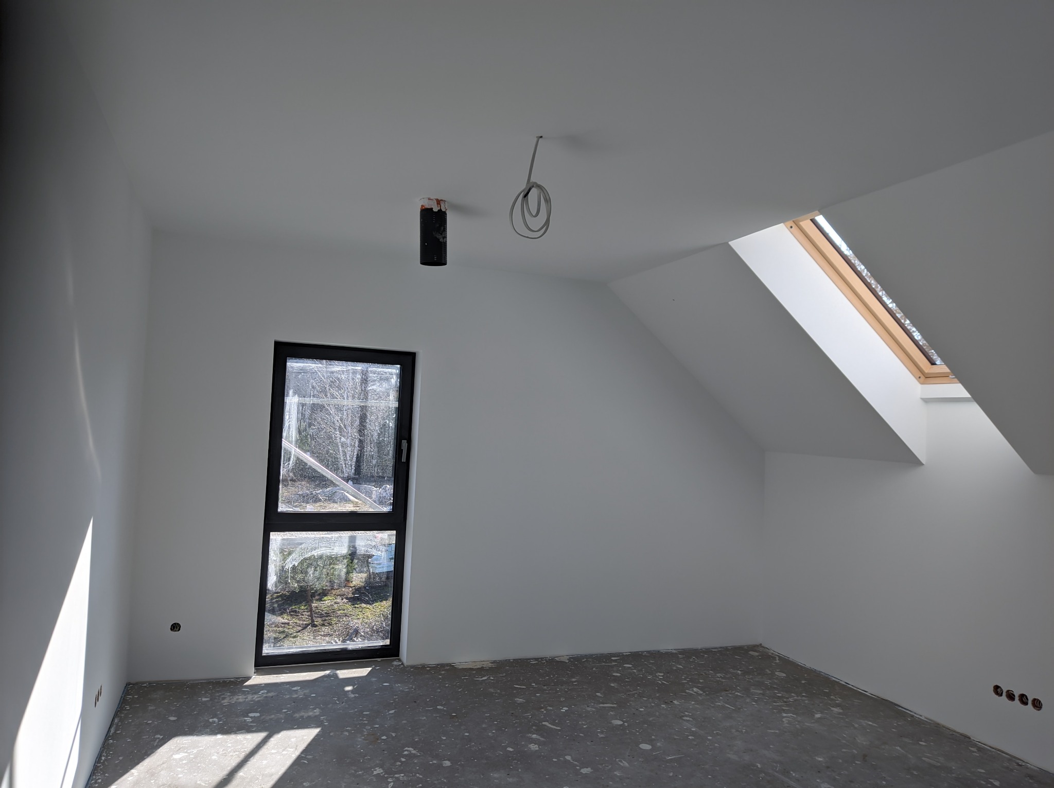 Poddasze w stanie deweloperskim z oknem dachowym i dużym oknem balkonowym, ściany pokryte świeżą białą farbą, widoczne wystające przewody elektryczne.