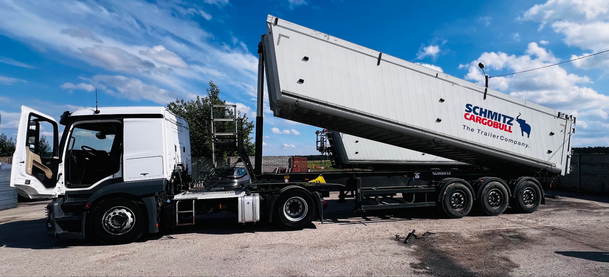 Biała ciężarówka z naczepą wywrotką Schmitz Cargobull z otwartą kabiną kierowcy, widoczna drabinka i logo firmy na naczepie, na tle błękitnego nieba z chmurami.