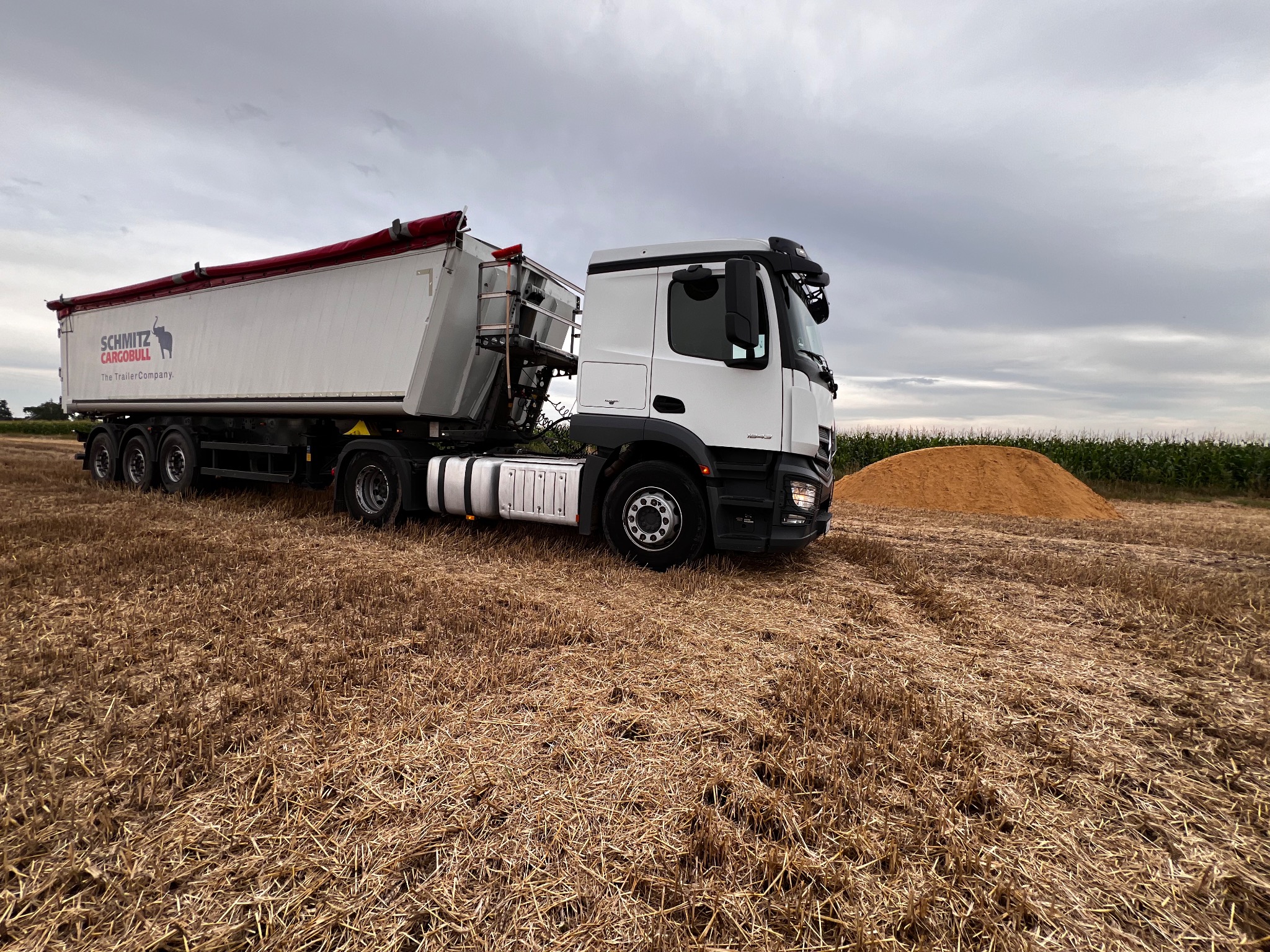Biała ciężarówka z naczepą Schmitz Cargobull na polu po żniwach, z widocznym stosem piasku w tle, pochmurne niebo.