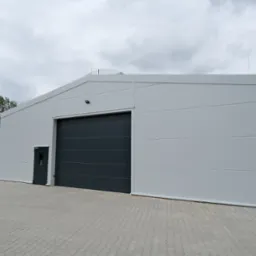 Gotowa biała hala z szarą bramą segmentową i drzwiami wejściowymi, widok z zewnątrz, na betonowej kostce, przy pochmurnym niebie.