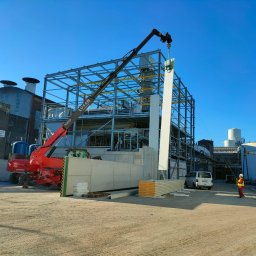 CRAFT CONSTRUCTION SPÓŁKA Z OGRANICZONĄ ODPOWIEDZIALNOŚCIĄ - Montaż płyty warstwowej na stalowej konstrukcji hali przemysłowej z użyciem dźwigu teleskopowego, pracownik w odblaskowej kamizelce kontroluje proces, w tle widoczne zbiorniki i budynki przemysłowe...