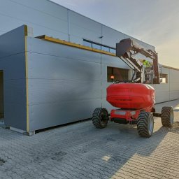 CRAFT CONSTRUCTION SPÓŁKA Z OGRANICZONĄ ODPOWIEDZIALNOŚCIĄ - Montaż szarej płyty warstwowej na ścianie hali przemysłowej z użyciem czerwonego podnośnika koszowego Manitou, w tle wschodzące słońce.