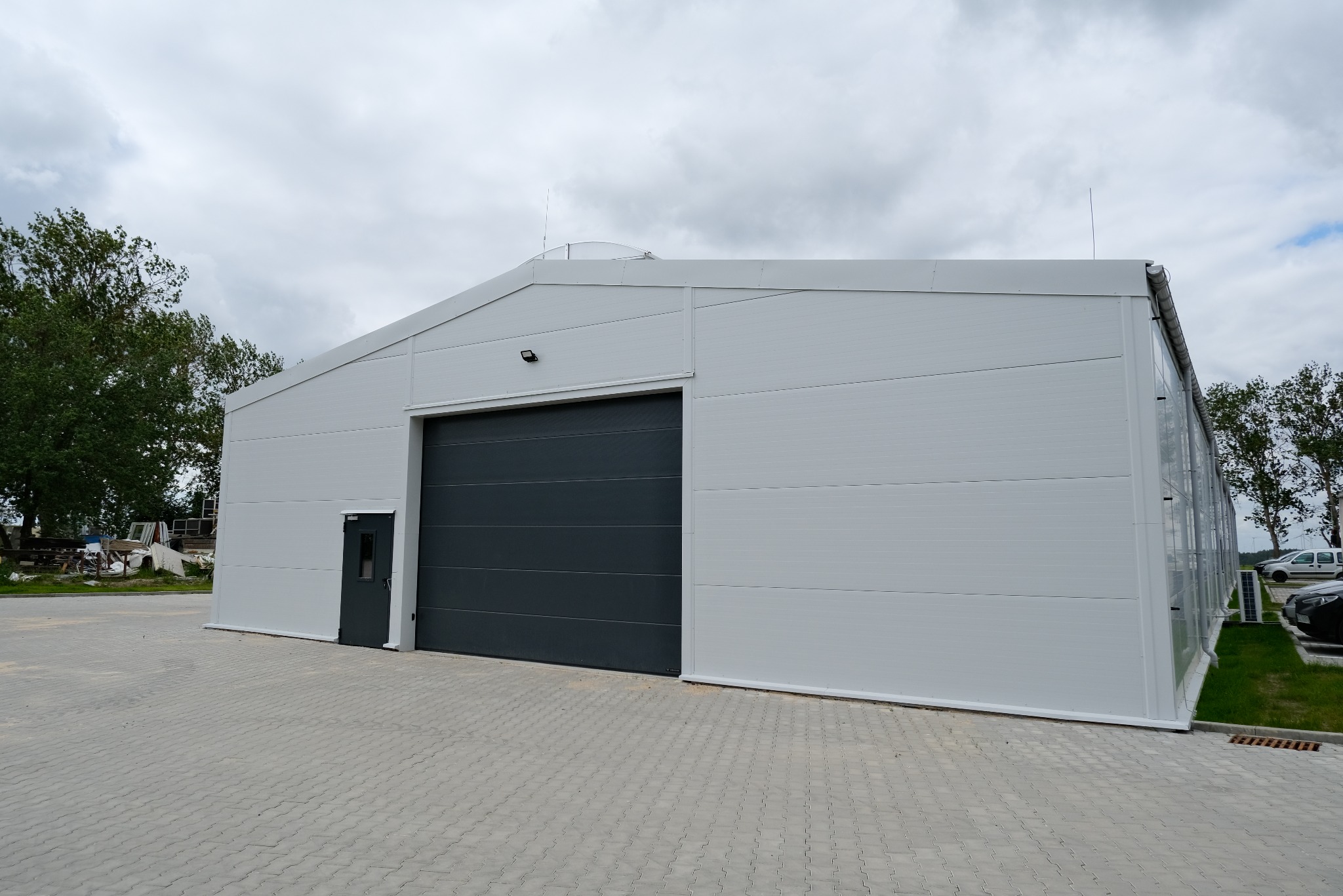 Gotowa biała hala z szarą bramą segmentową i drzwiami wejściowymi, widok z zewnątrz, na betonowej kostce, przy pochmurnym niebie.