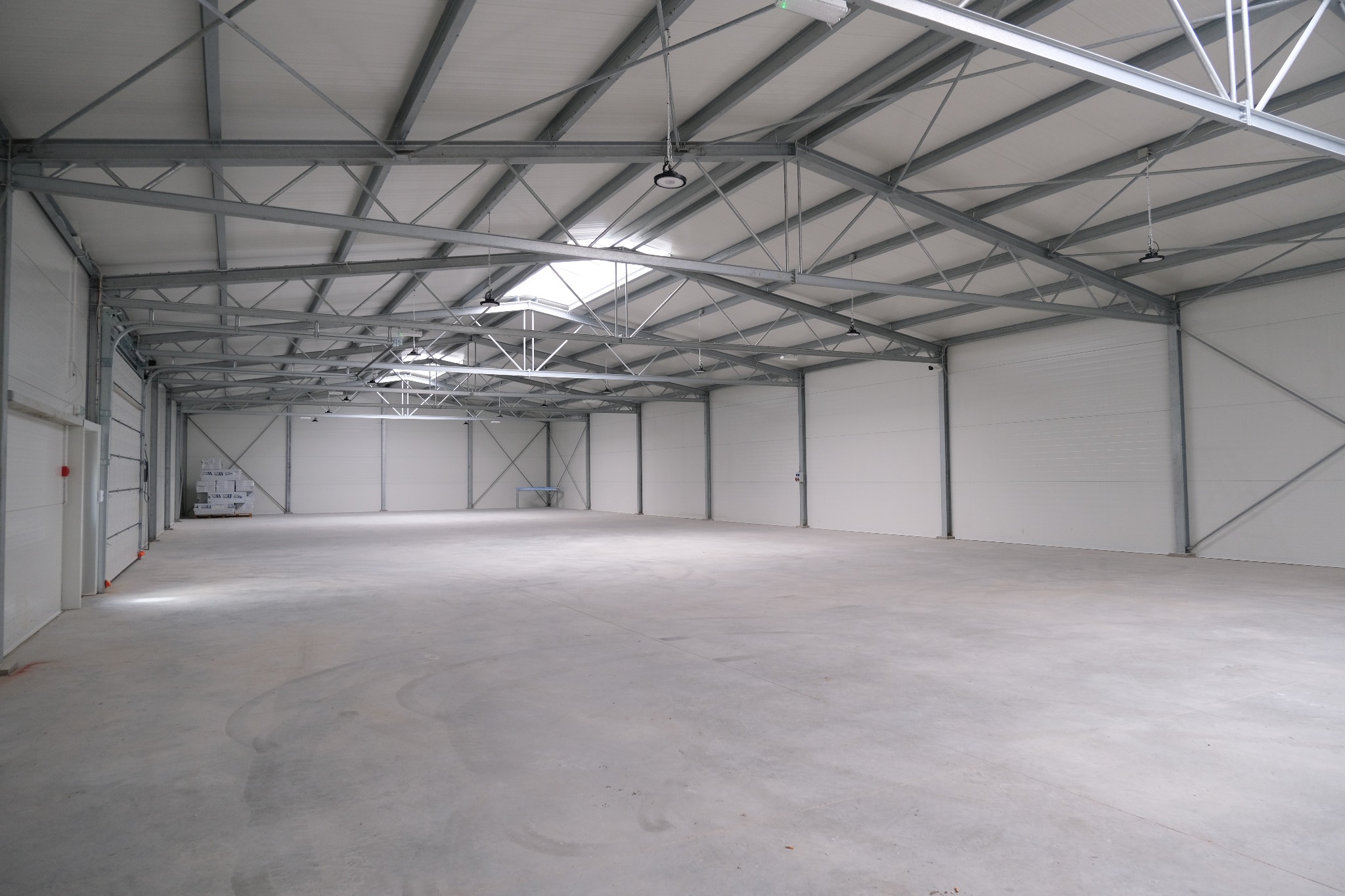 Wnętrze nowo wybudowanej hali z widoczną stalową konstrukcją dachu, białymi płytami warstwowymi na ścianach i betonową posadzką. W tle widoczny mały stolik.
