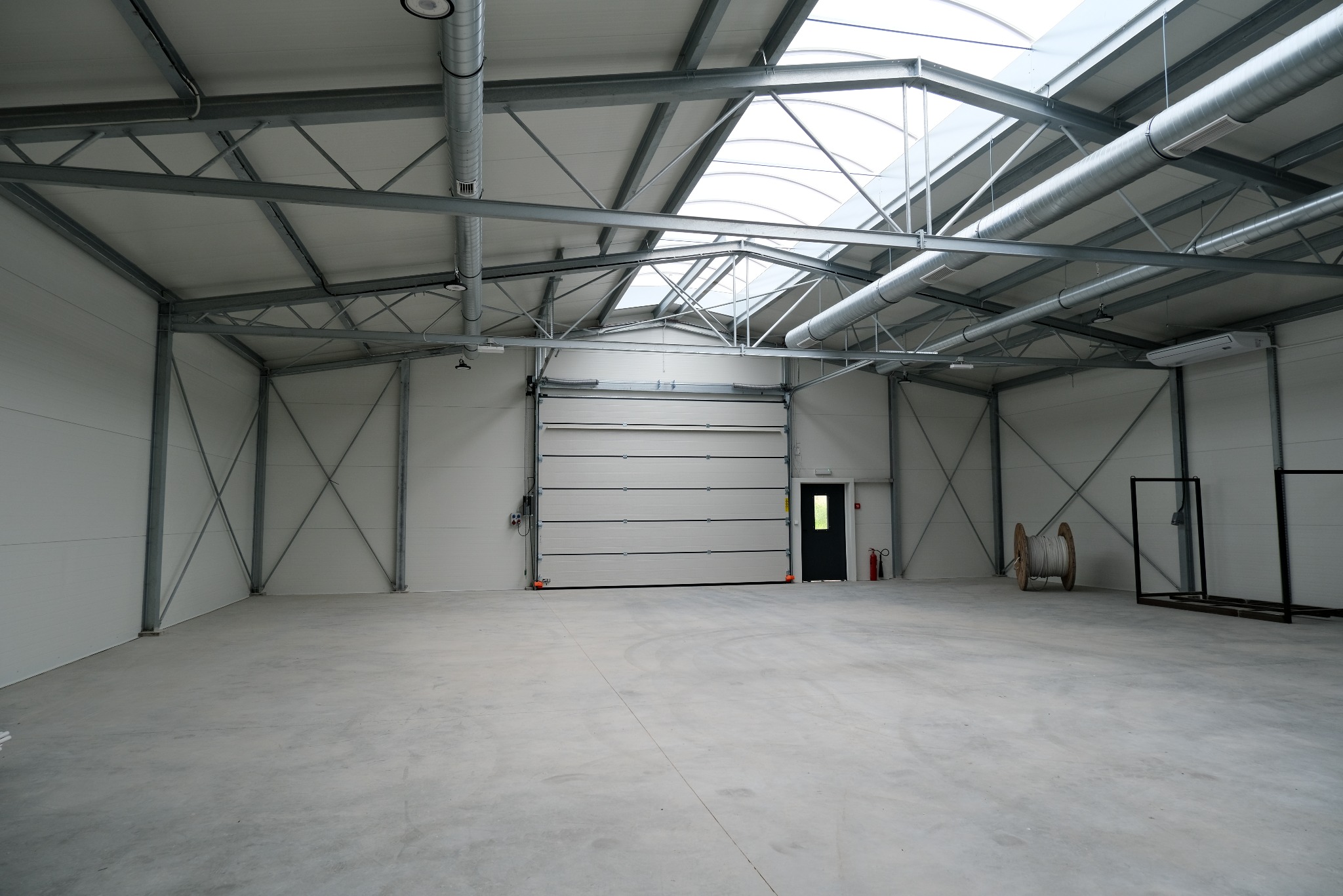Wnętrze nowo wybudowanej hali magazynowej z szarą stalową konstrukcją, białymi ścianami z płyt warstwowych i betonową posadzką. Widoczna brama segmentowa, świetliki dachowe i zwój kabla...