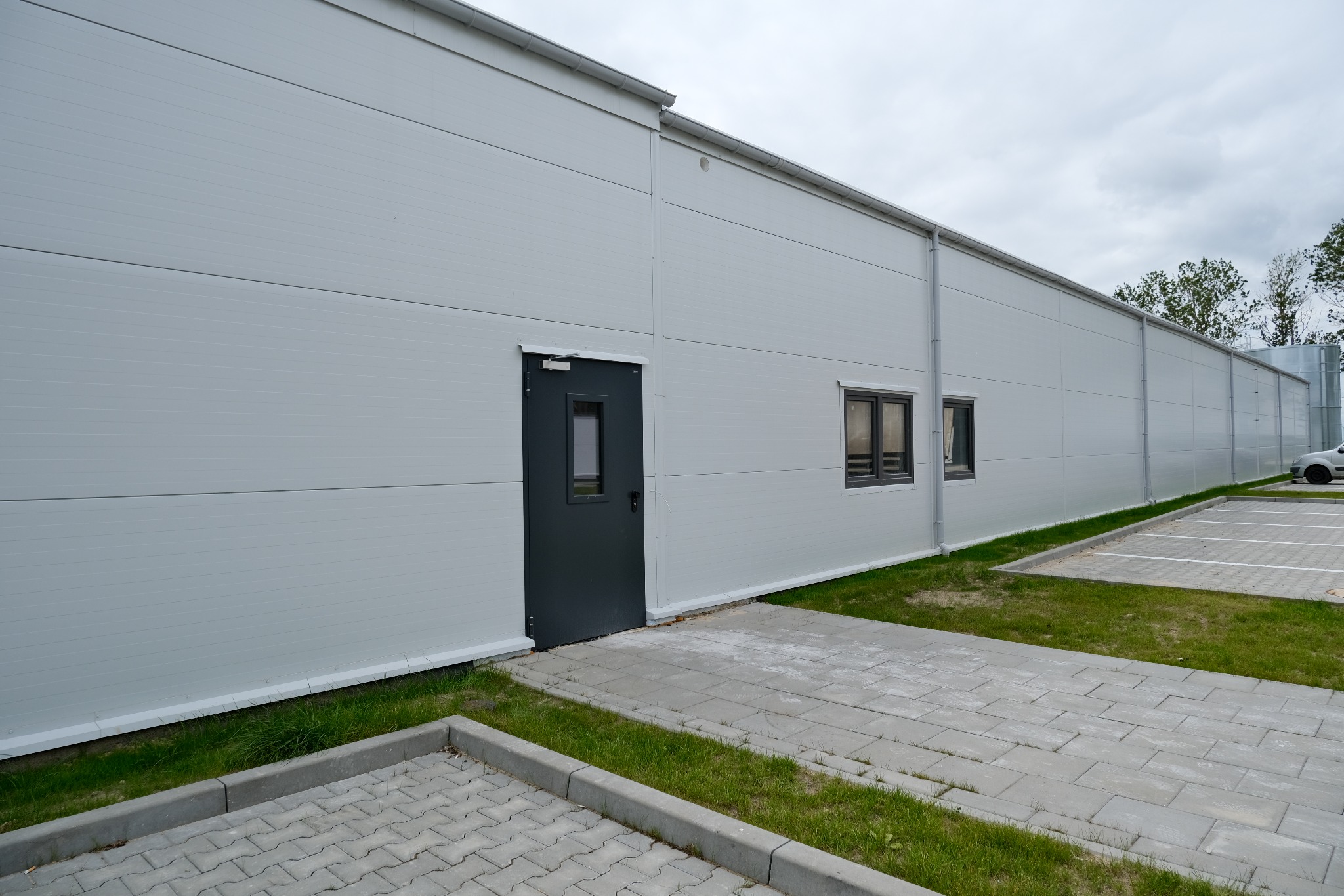 Zewnętrzny widok nowo wybudowanej hali z białych płyt warstwowych, z szarą stolarką okienną i drzwiami, oraz fragmentem parkingu z wyznaczonymi miejscami postojowymi.
