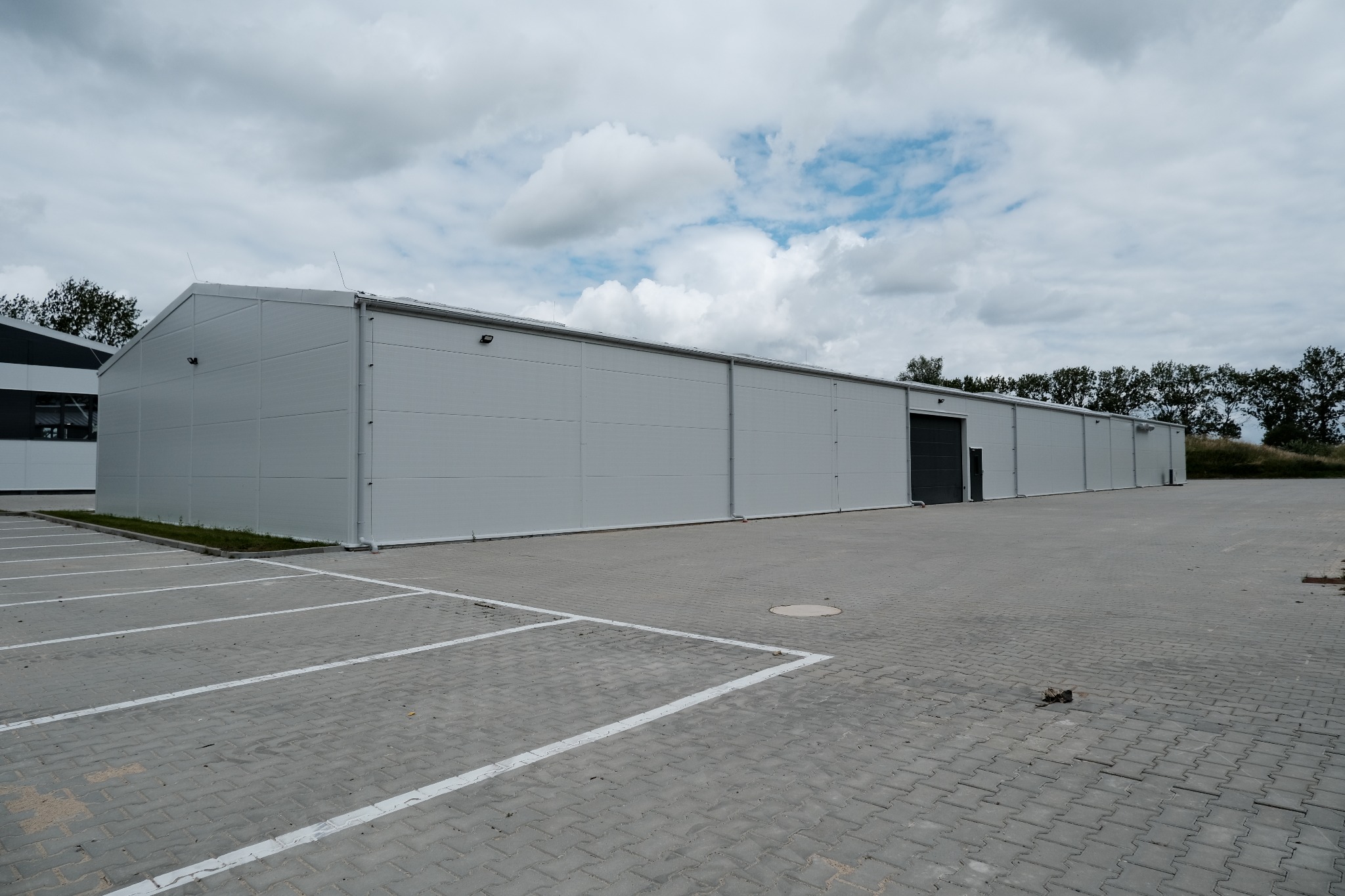 Gotowa hala magazynowa z szarej płyty warstwowej, widok z zewnątrz na parking z wyznaczonymi miejscami postojowymi i pochmurne niebo.