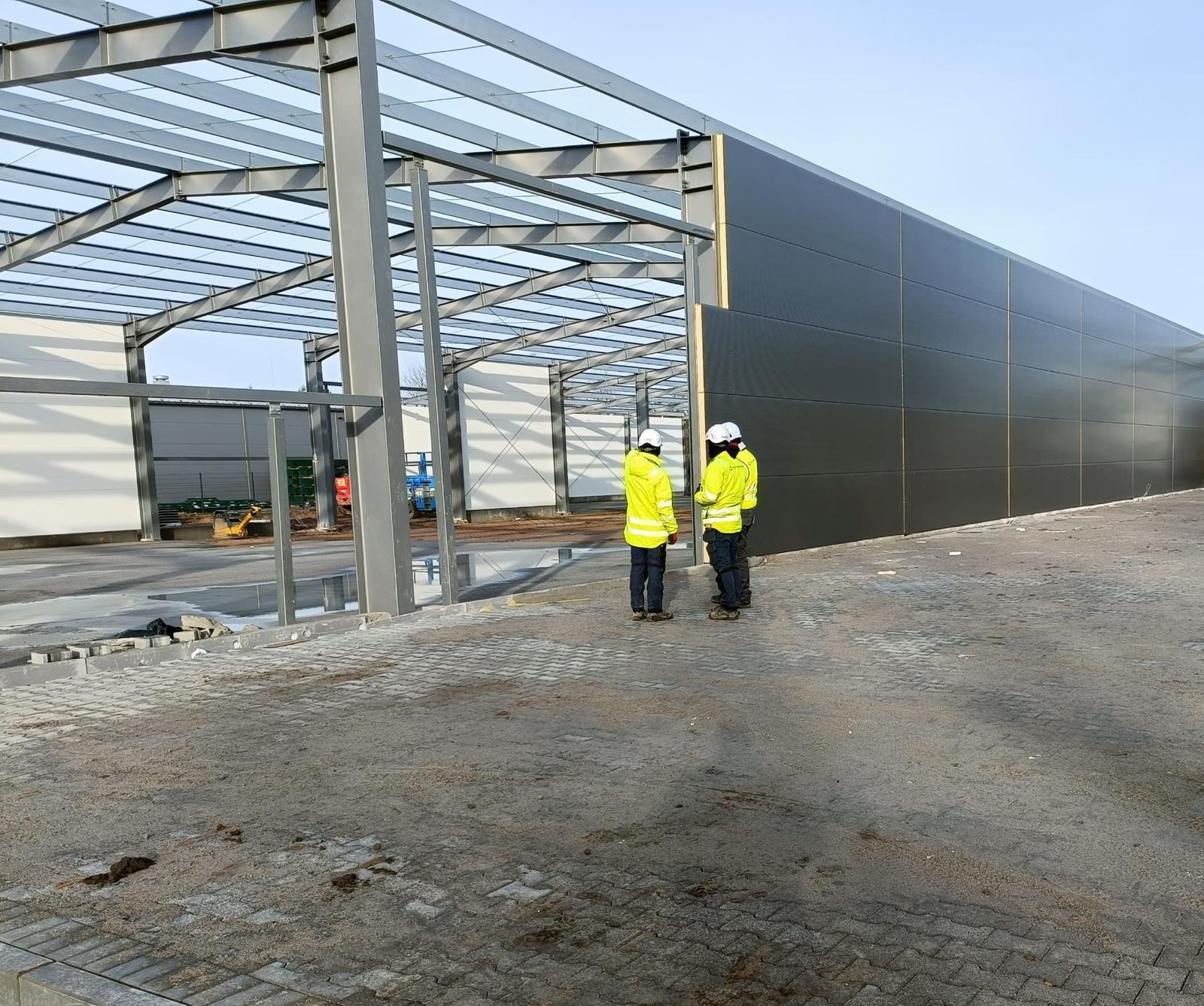 Montaż paneli warstwowych na stalowej konstrukcji hali, widoczni dwaj pracownicy w żółtych kurtkach i białych kaskach, teren placu budowy z kostki brukowej.