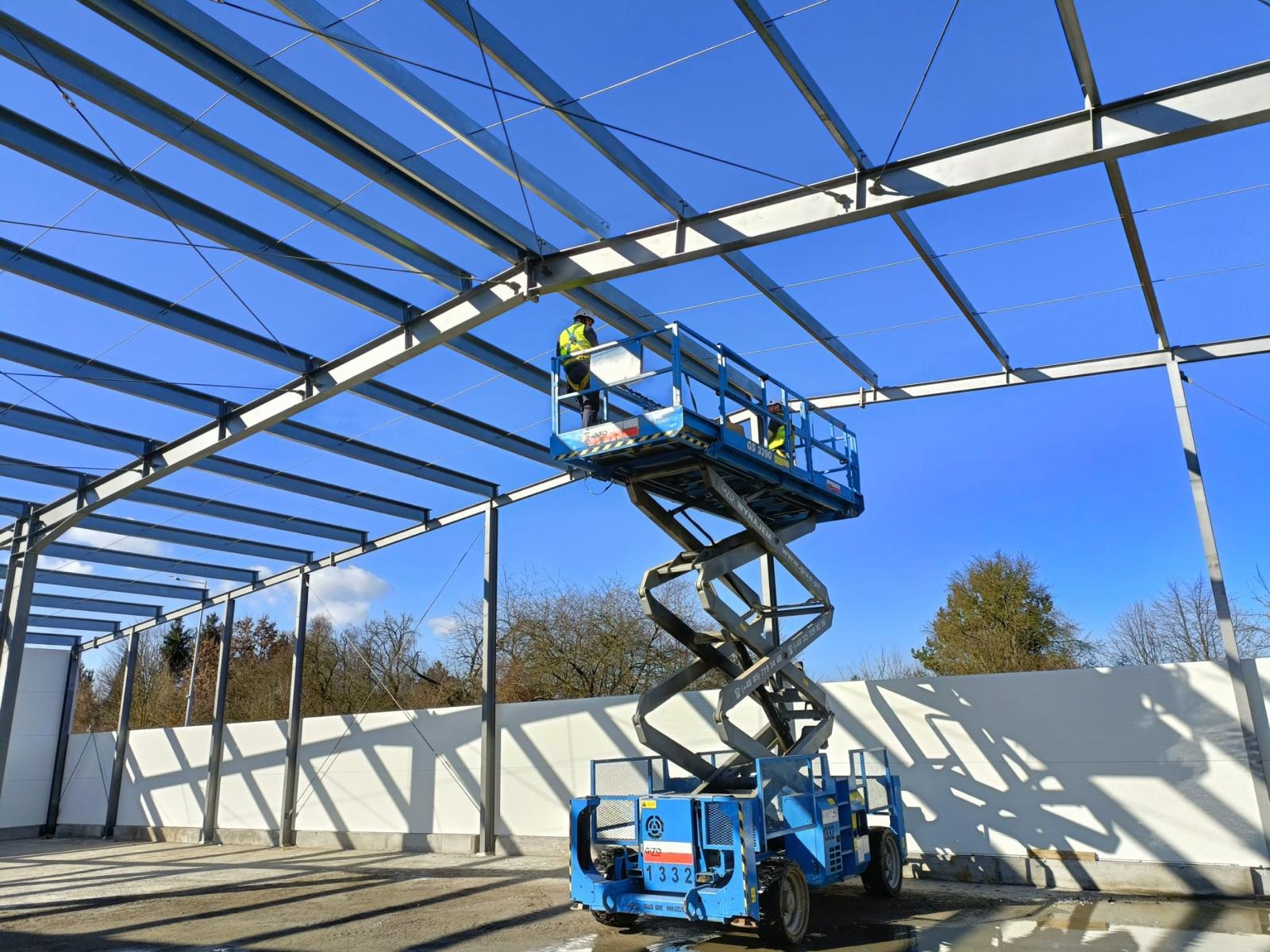 Dwóch pracowników w żółtych kamizelkach, na niebieskim podnośniku nożycowym, montuje stalową konstrukcję hali na tle błękitnego nieba. Widoczne elementy konstrukcyjne oraz częściowo zamontowane...